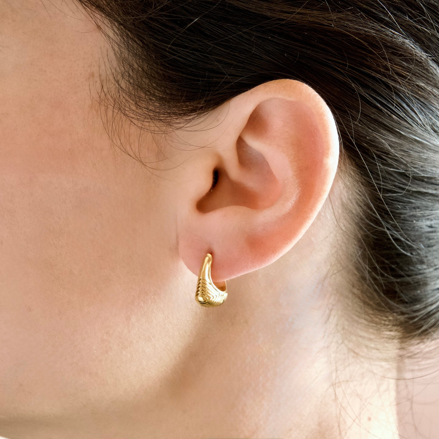 A close-up of a person's ear wearing a small textured gold hoop earring.