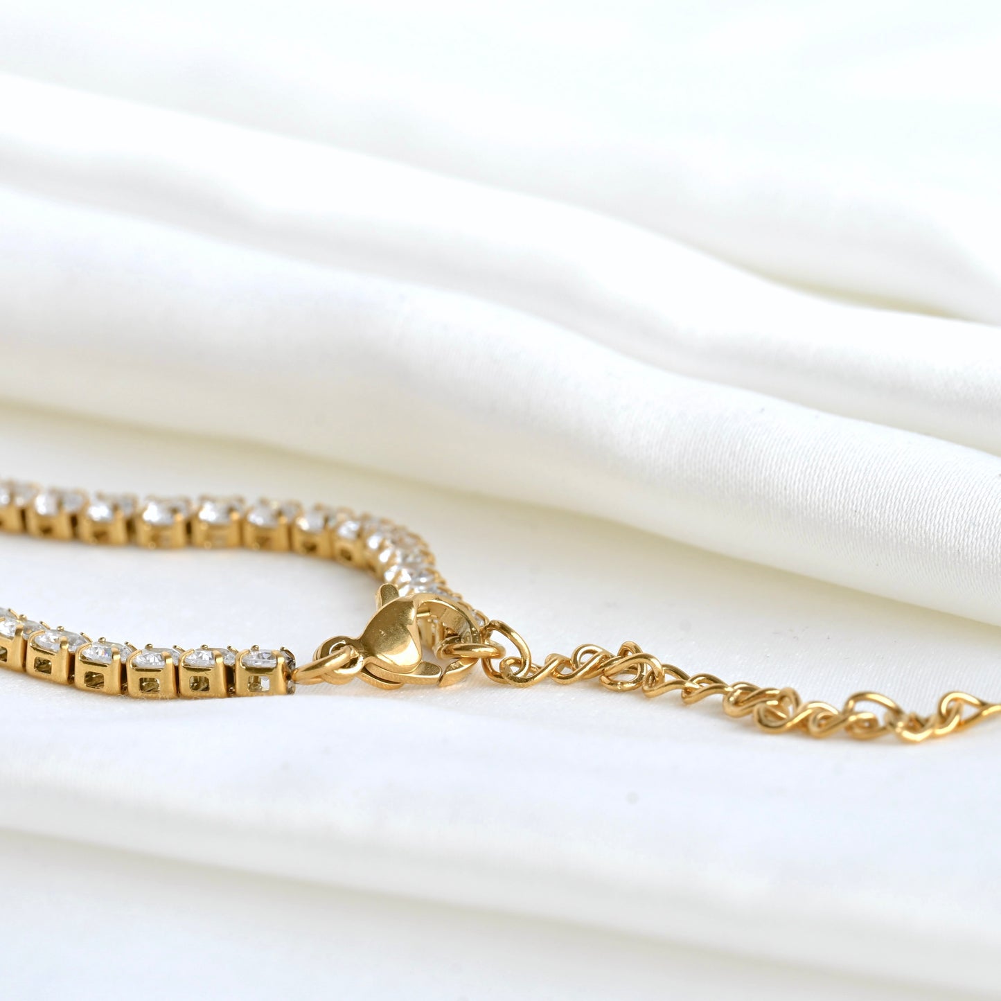 Gold tennis bracelet, close-up view of its lobster claw clasp and extender chain on a white fabric background