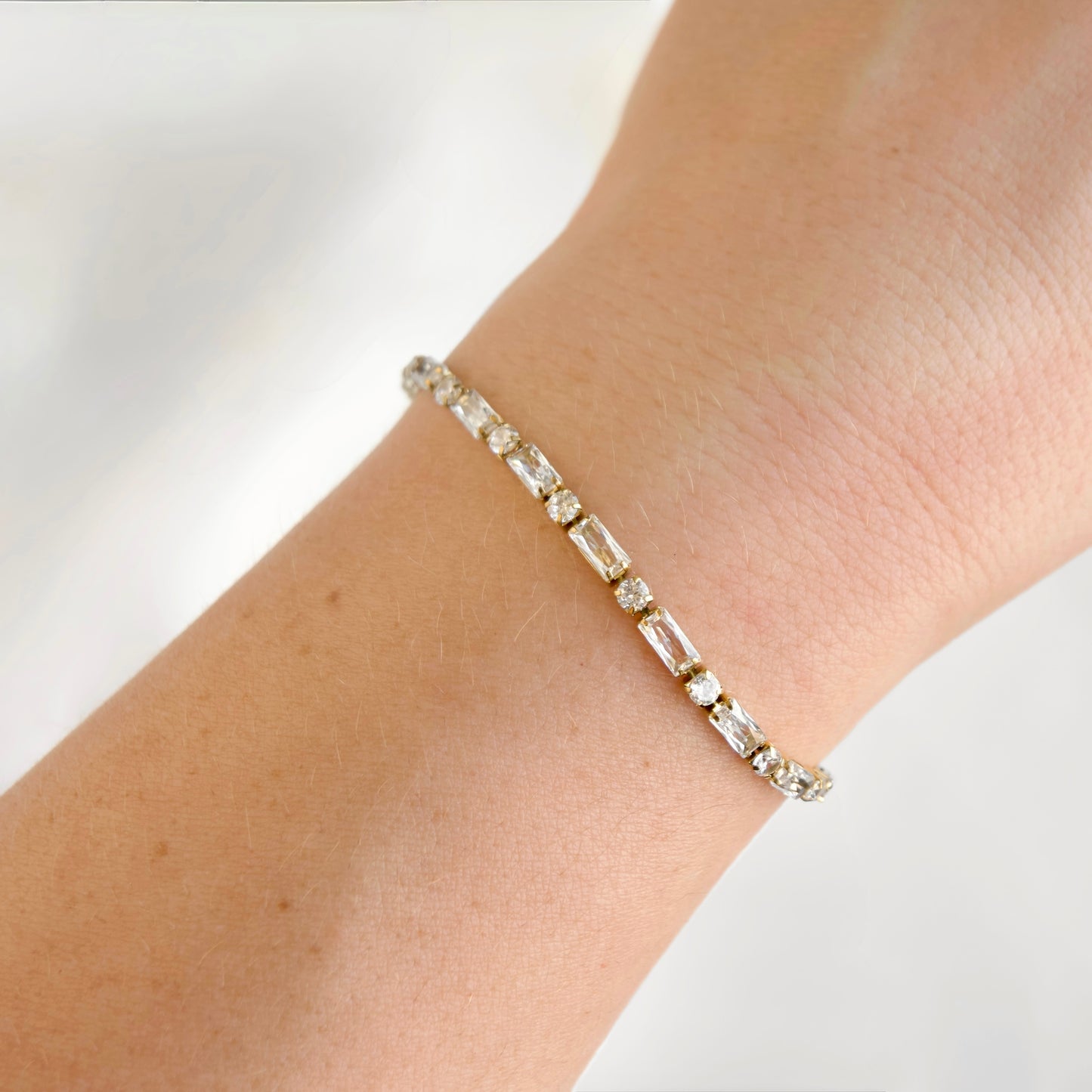 A delicate gold tennis bracelet with baguette-cut clear stones worn on a person's wrist against a soft, light background.