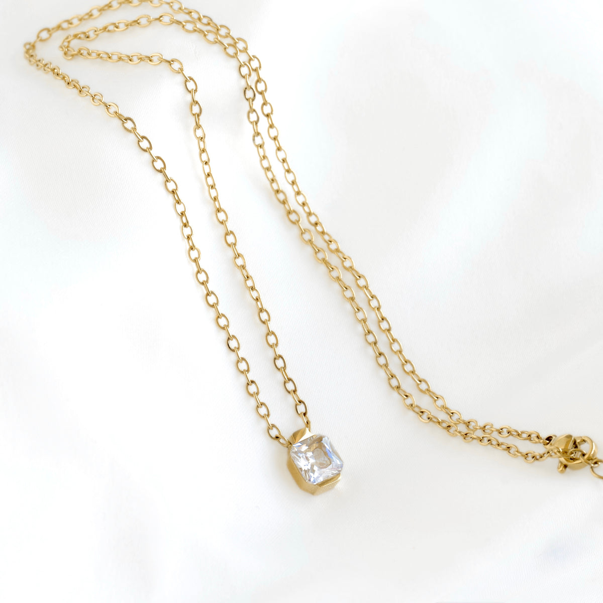 A delicate gold chain necklace with a bezel-set clear rectangular gemstone pendant, displayed on a soft white fabric background.