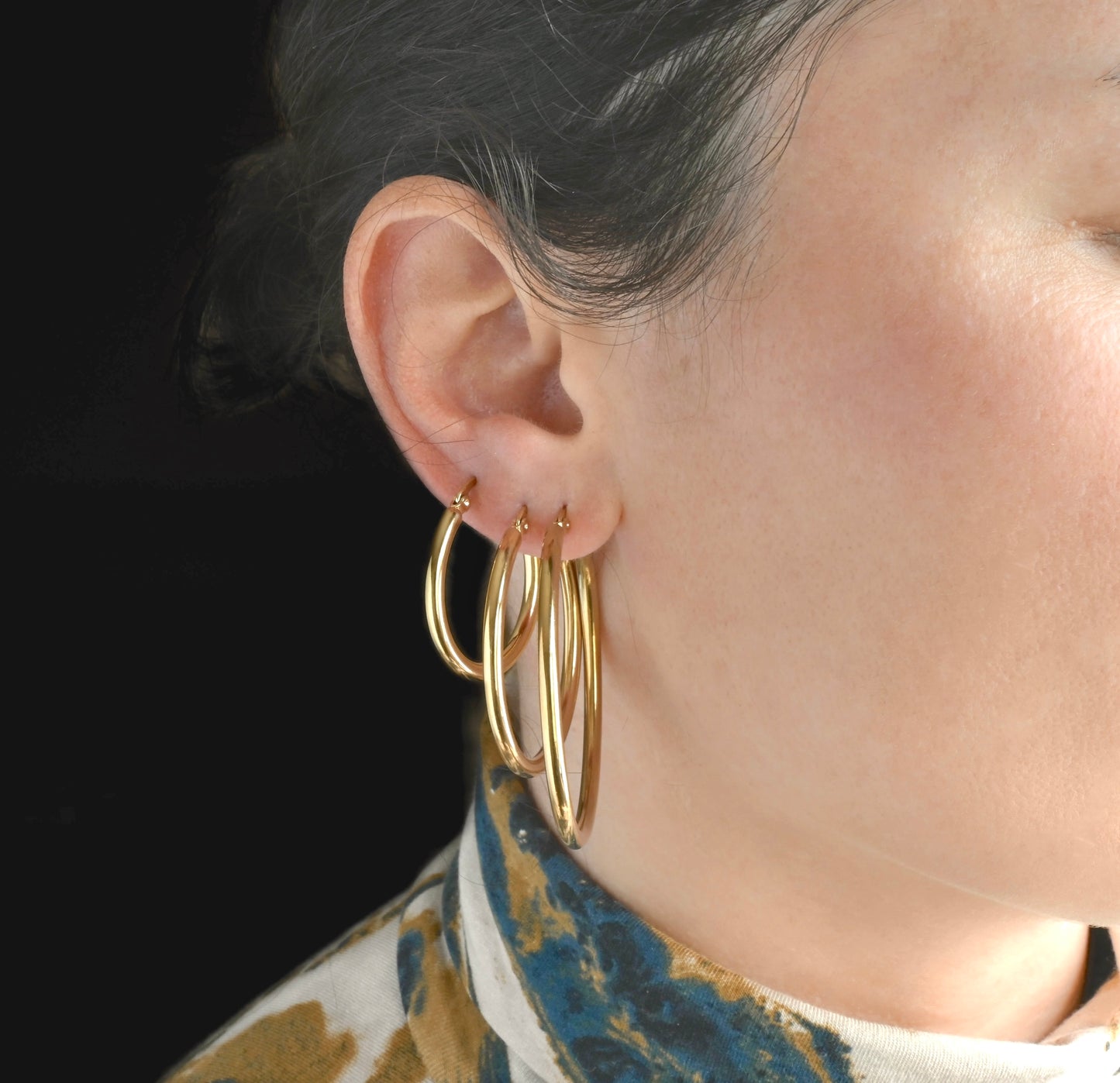 Close-up front view of a person wearing stacked gold tunnel hoop earrings in 30mm, 40mm and 50mm, with a dark background