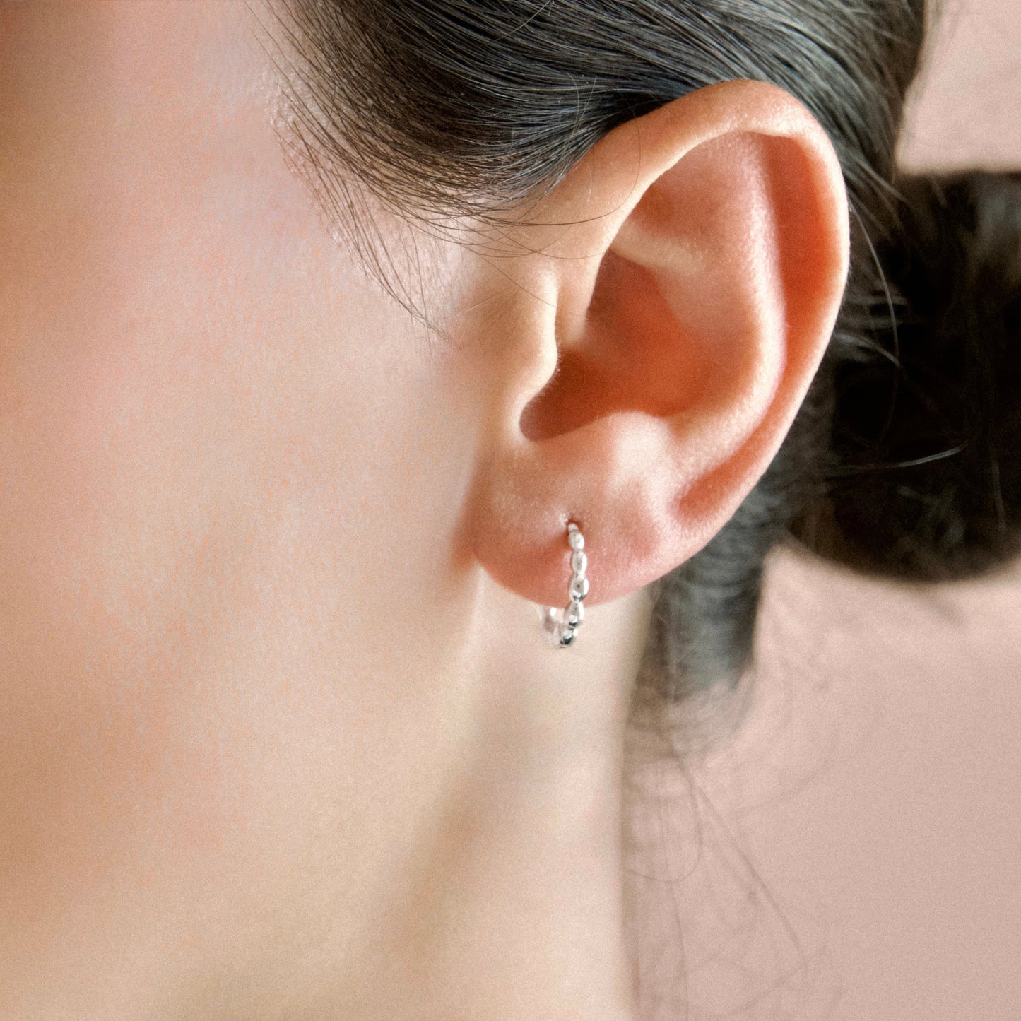 A close-up of a person's ear wearing a small silver huggie hoop earring  against a soft beige background.