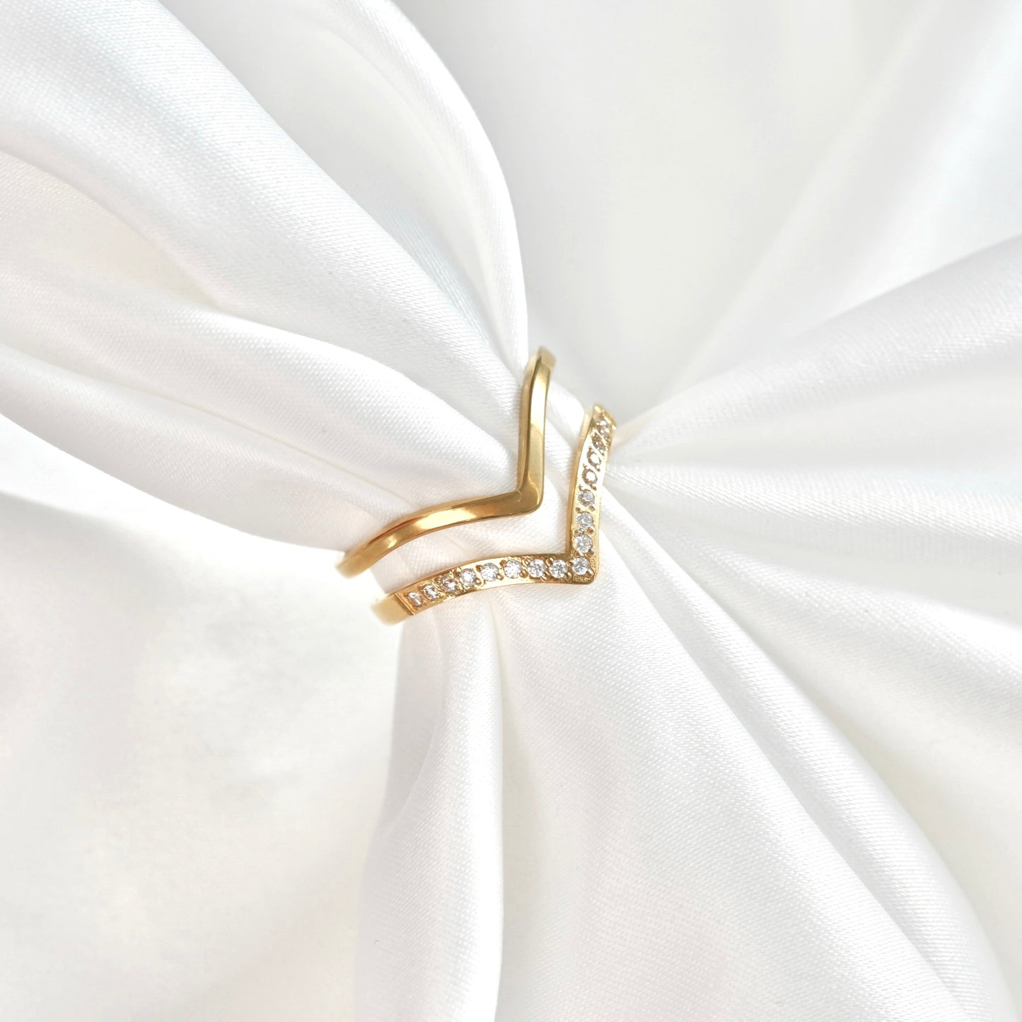 A gold V-shaped ring with a row of clear crystals on one side, displayed on a white satin fabric background.