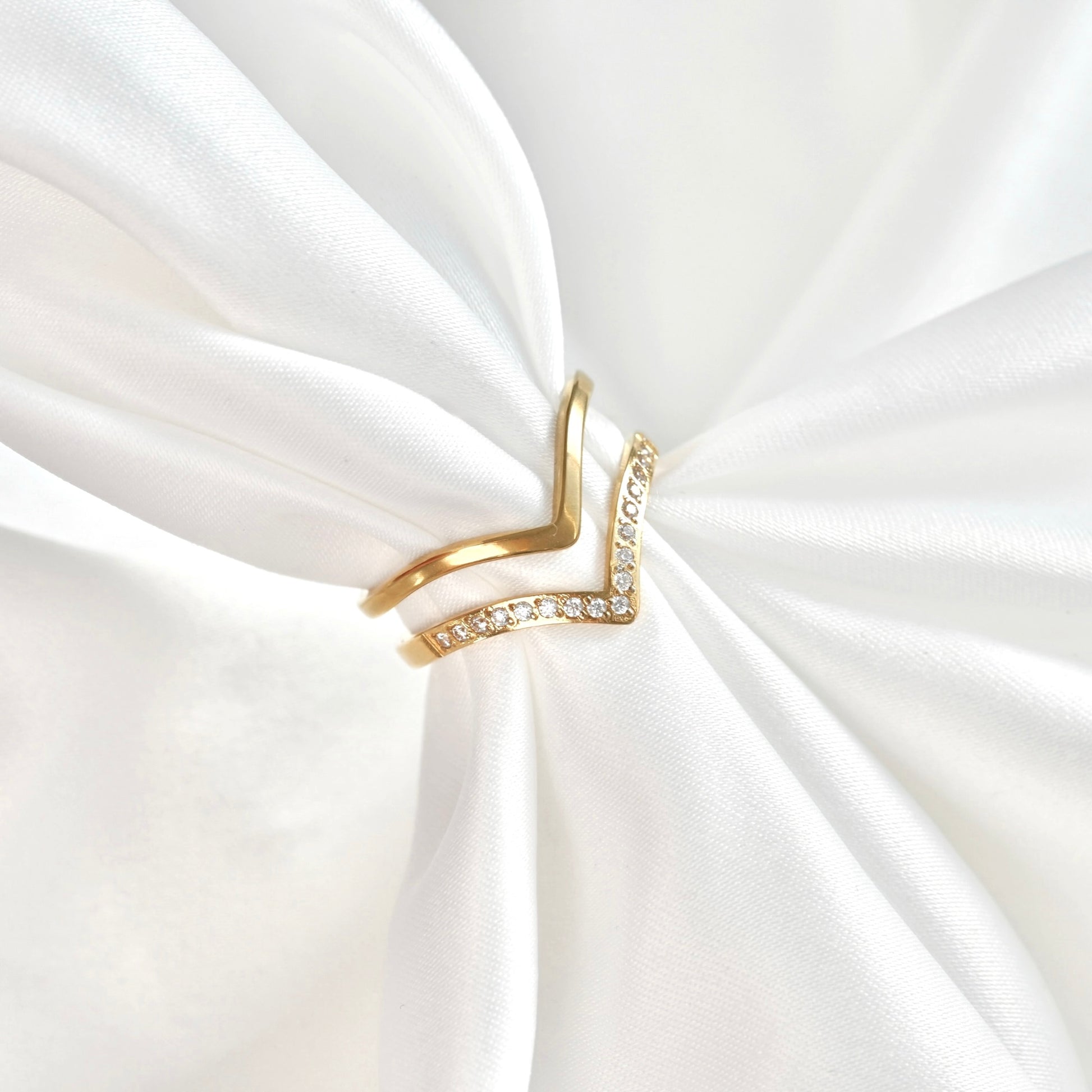 A gold V-shaped ring with a row of clear crystals on one side, displayed on a white satin fabric background.