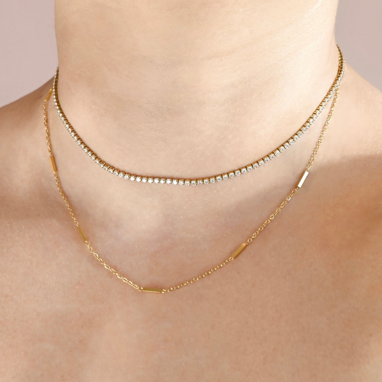 A close-up of a model's neck and upper chest wearing two layered gold necklaces: one delicate tennis-style chain with small clear stones and one longer chain with polished gold bar accents.