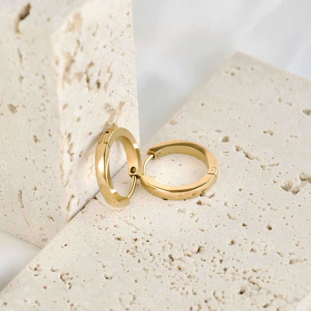 A pair of small polished gold hoop earrings, resting on and against beige stone.