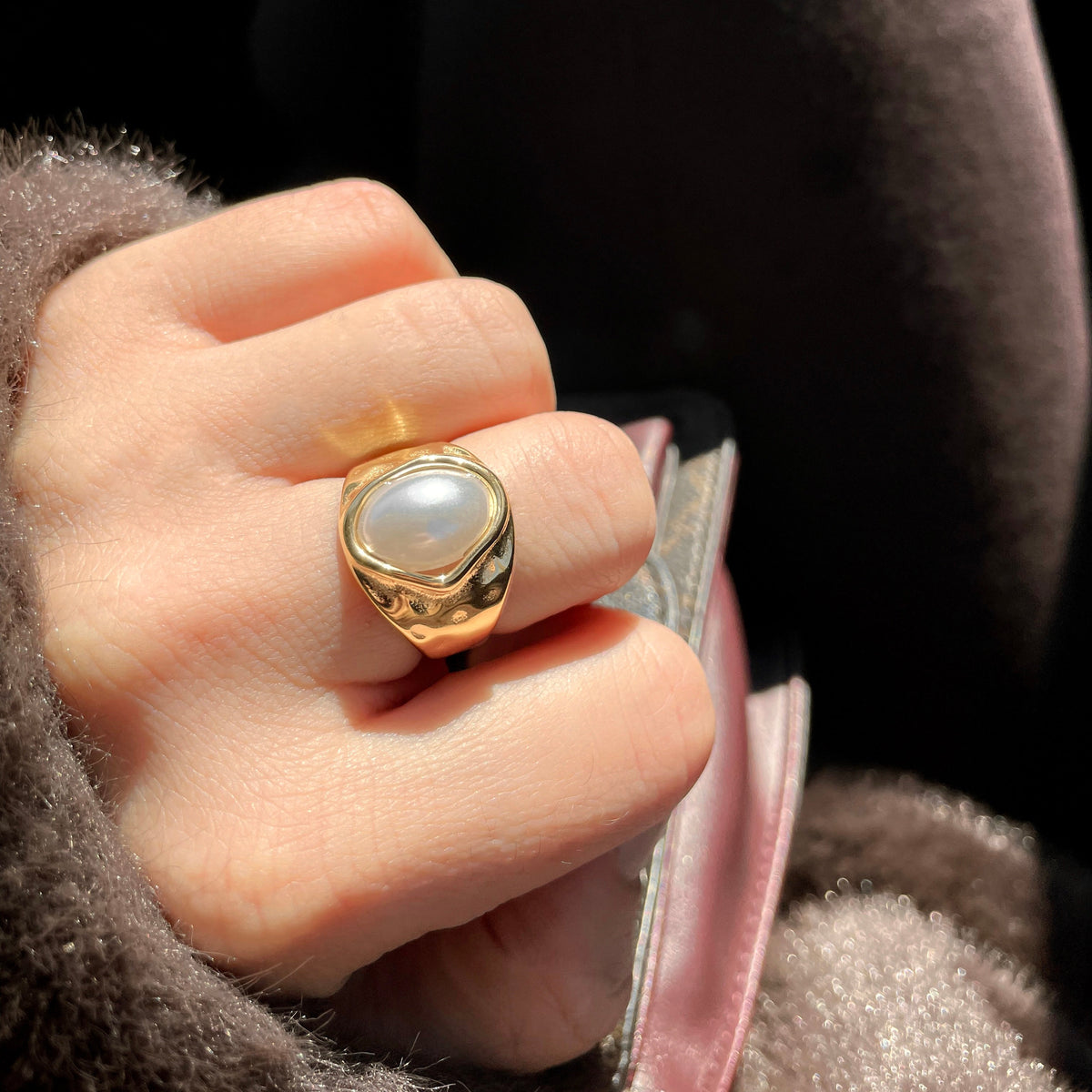 A gold signet ring with a round white pearl-like stone worn on a hand, set against a dark brown textured background.