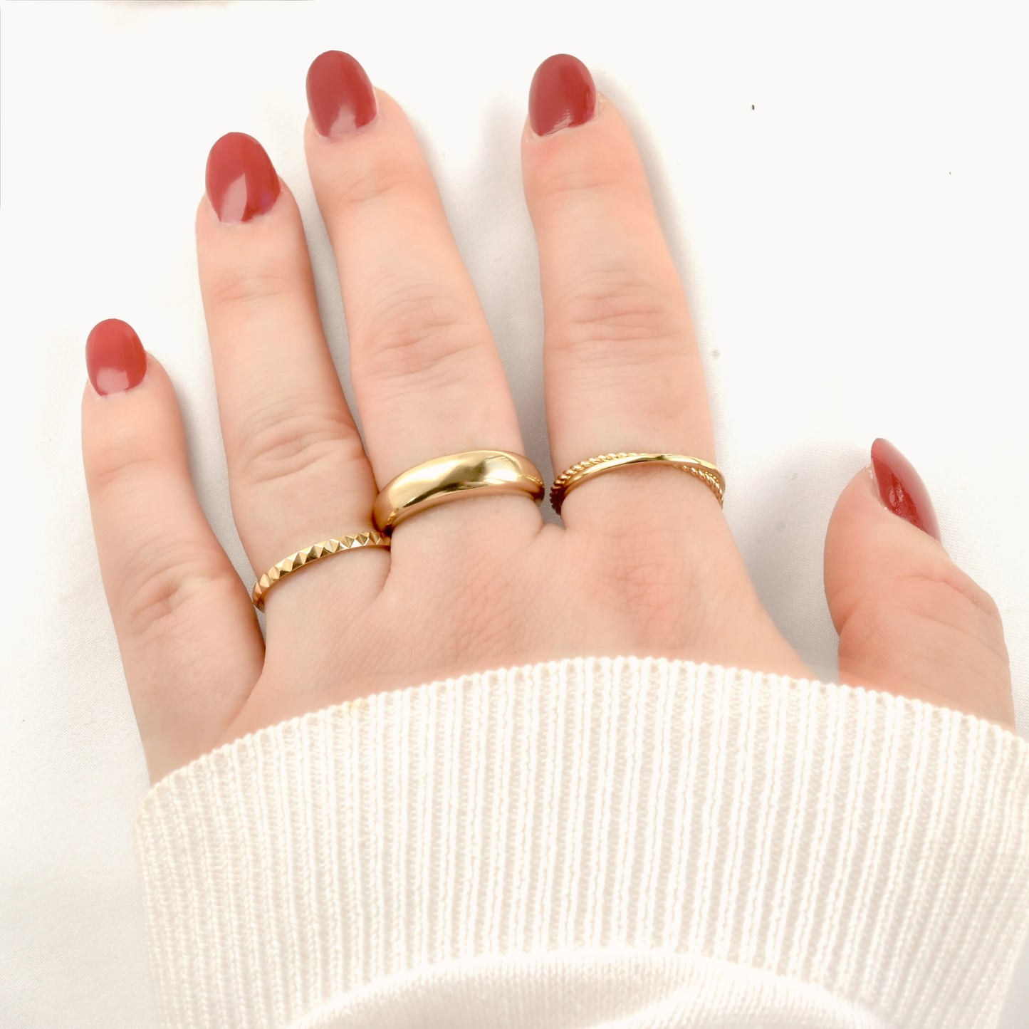 Hand wearing three gold rings, a slim pyramid design, chunky dome ring and interlocking rings  on a white background
