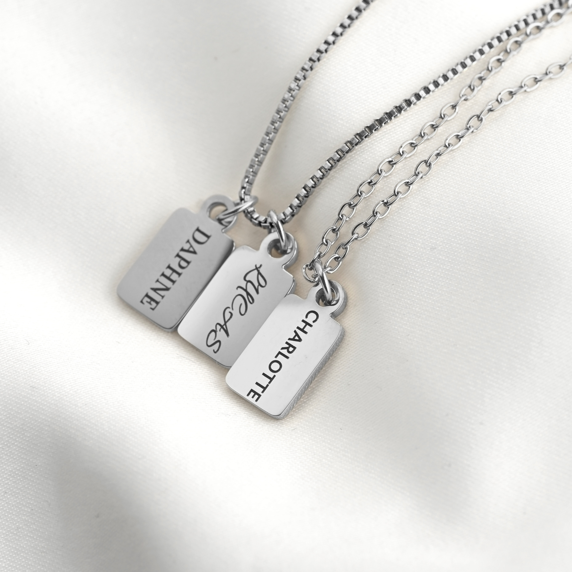 Three silver rectangular nameplates on matching chains, engraved with the names DAPHNE, LUCAS, and CHARLOTTE, displayed on a soft white fabric background.