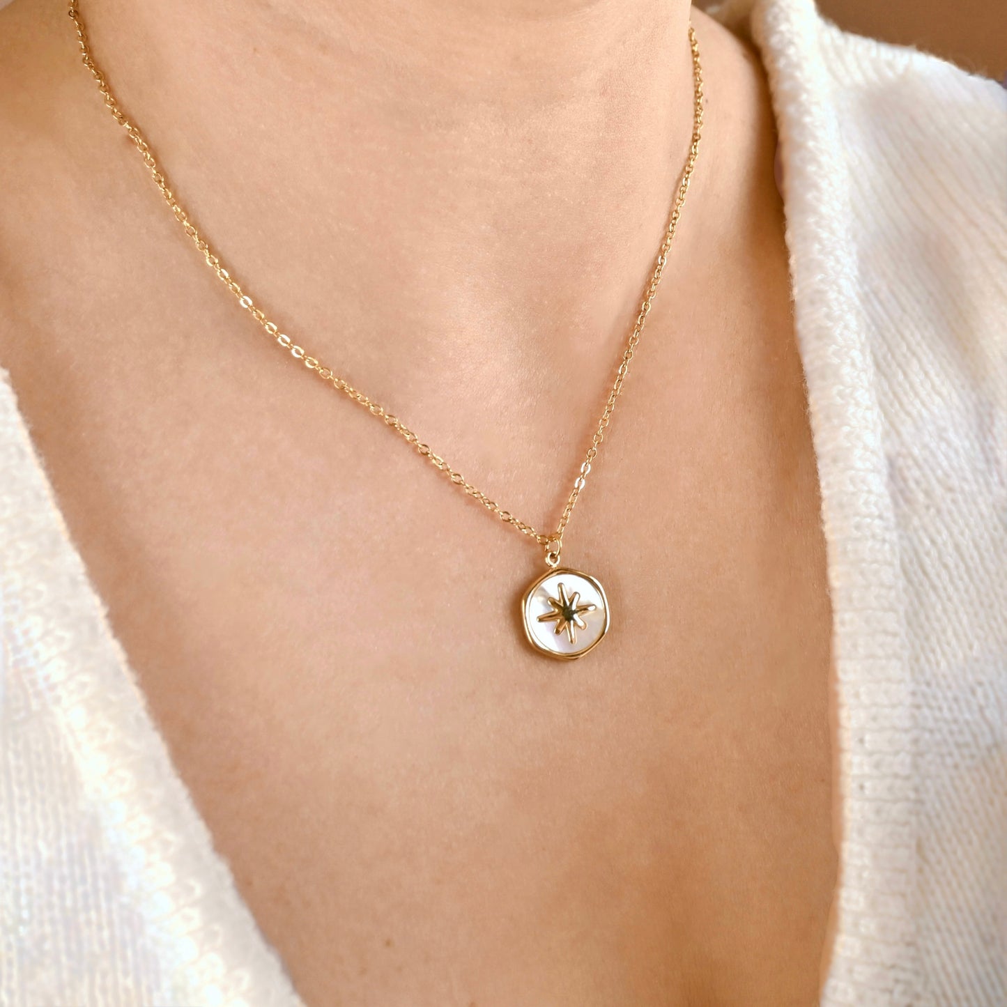 A close-up of a person wearing a delicate gold chain necklace with a small round pendant featuring a white inlay and a gold compass rose design.