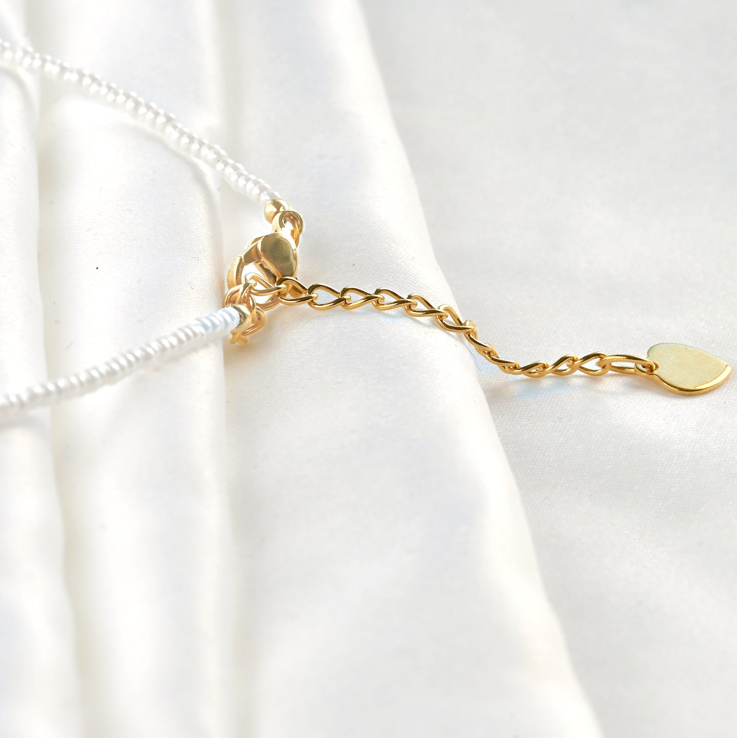 Close up view of the stainless steel gold clasp, extender and a tiny flat heart charm on white seed bead necklace on a white background