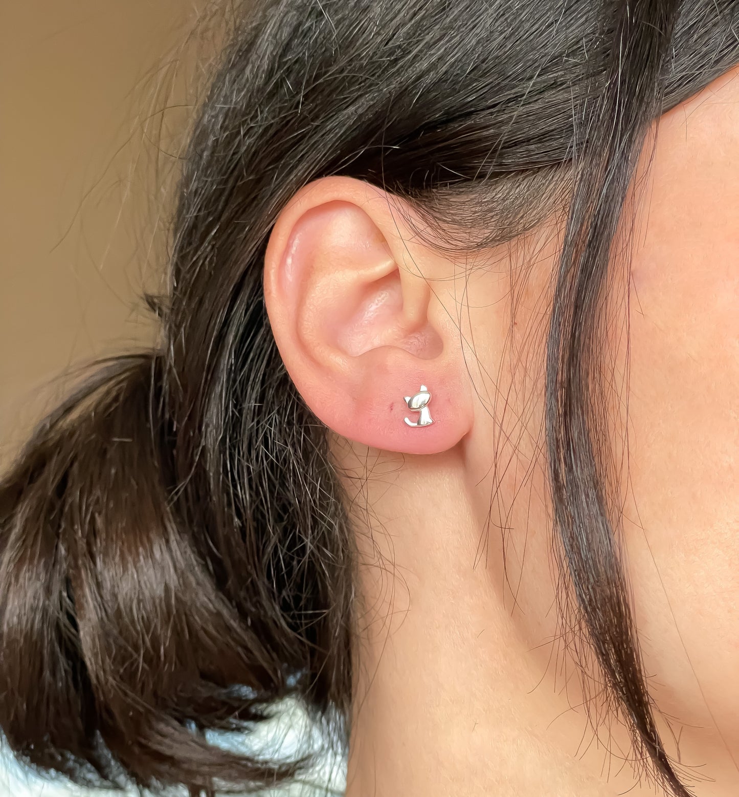 Close-up of an ear wearing small silver kitten stud earrings with a neutral background