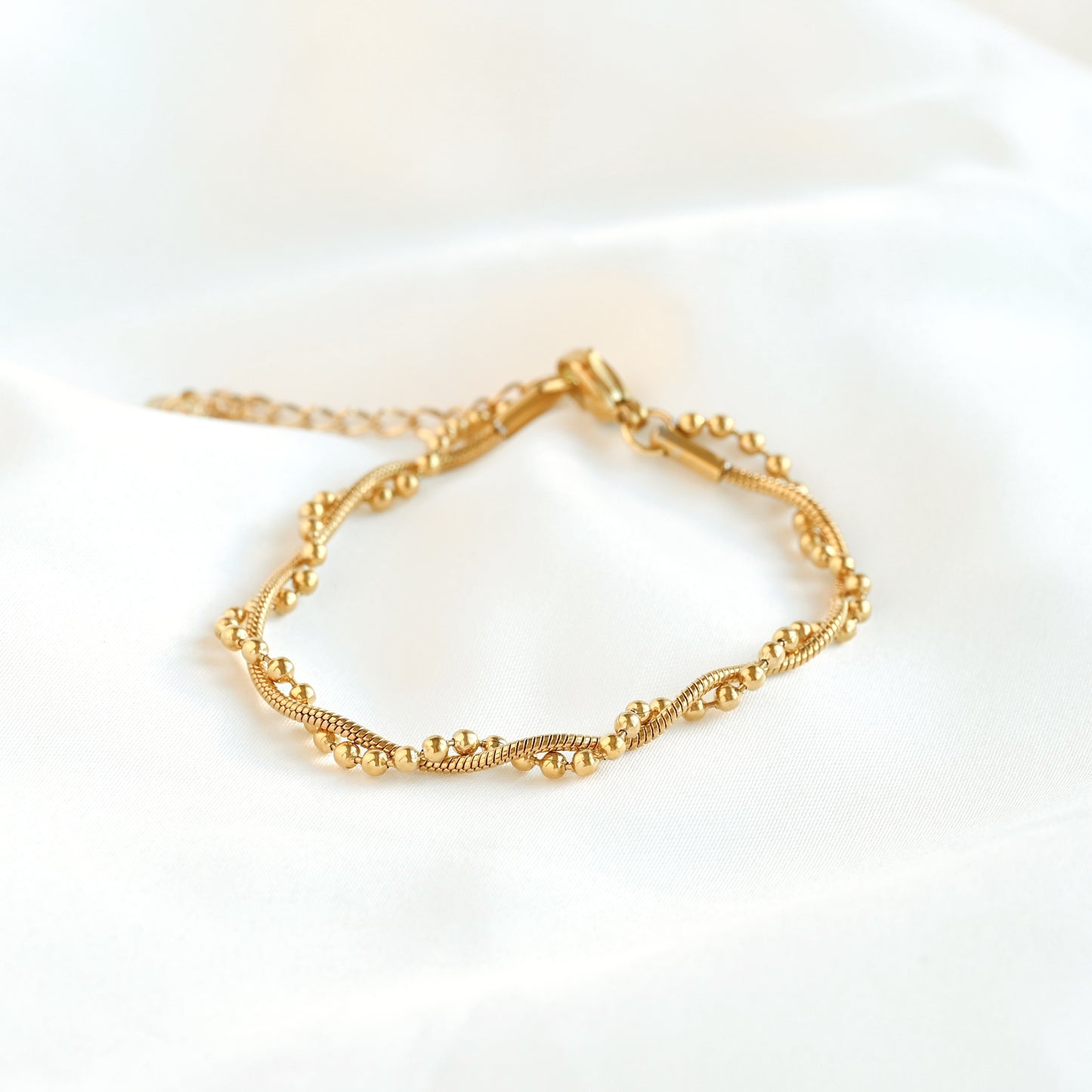 A delicate gold chain bracelet with small bead-like links and a lobster clasp, displayed on a soft white fabric background.