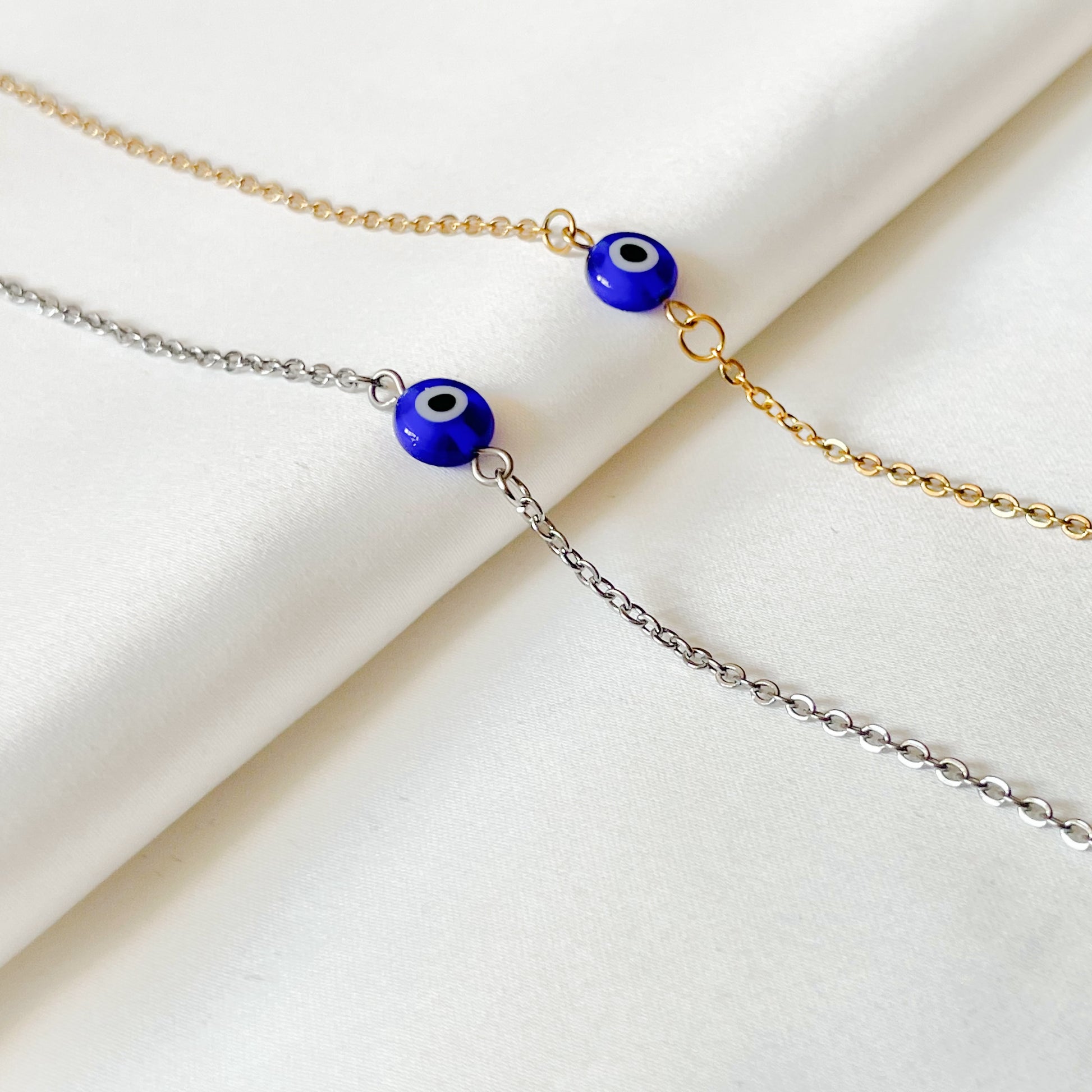 Two ankle bracelets one gold and one silver with blue evil eye beads on a beige fabric background
