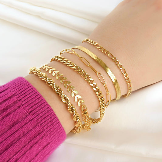 Gold bracelet stack on a person's wrist with a pink sleeve, with chunky rope, leaf, cuban, paperclip, herringbone and figaro chains.