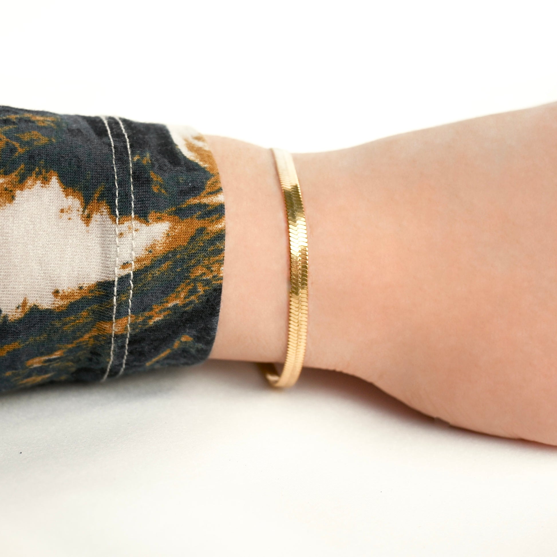 Wide gold herringbone bracelet on a wrist with a patterned sleeve against a white background
