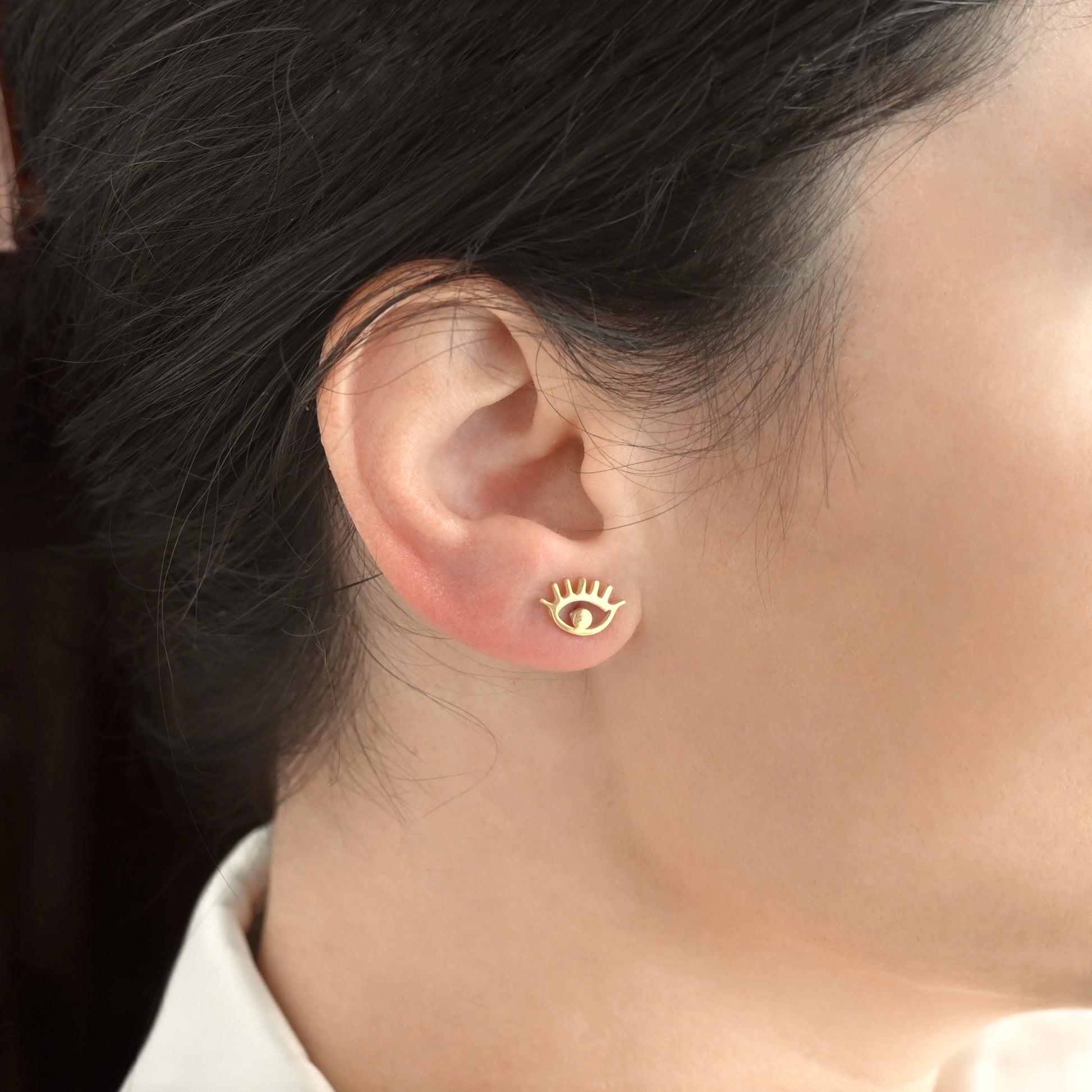 Gold eye-shaped stud earrings with lashes worn by a person with dark hair.