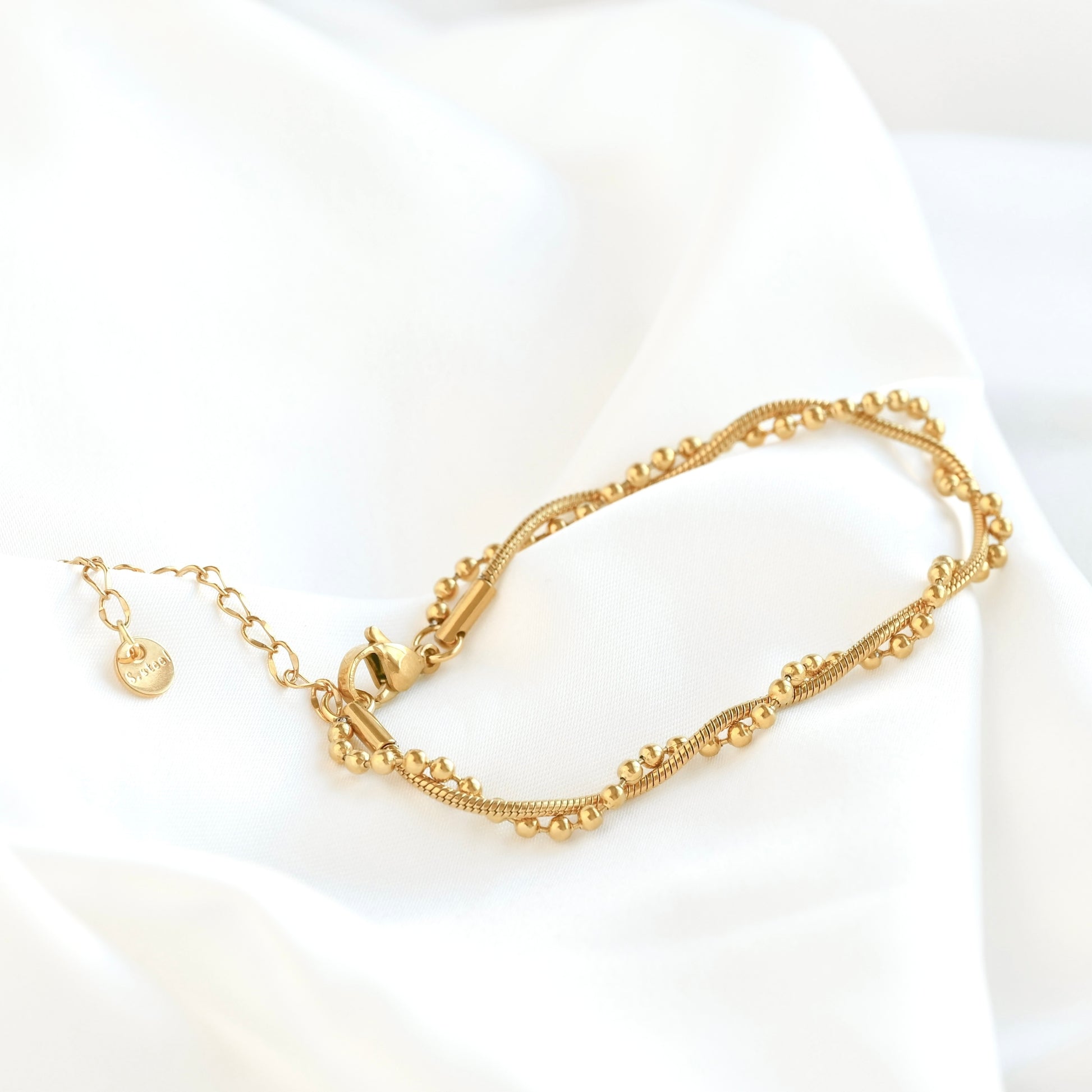 A delicate gold chain bracelet draped on a soft white fabric background. The bracelet features a textured snake-style chain with small bead accents along the top and bottom edges, a clasp closure, and an extender chain with a small round charm.