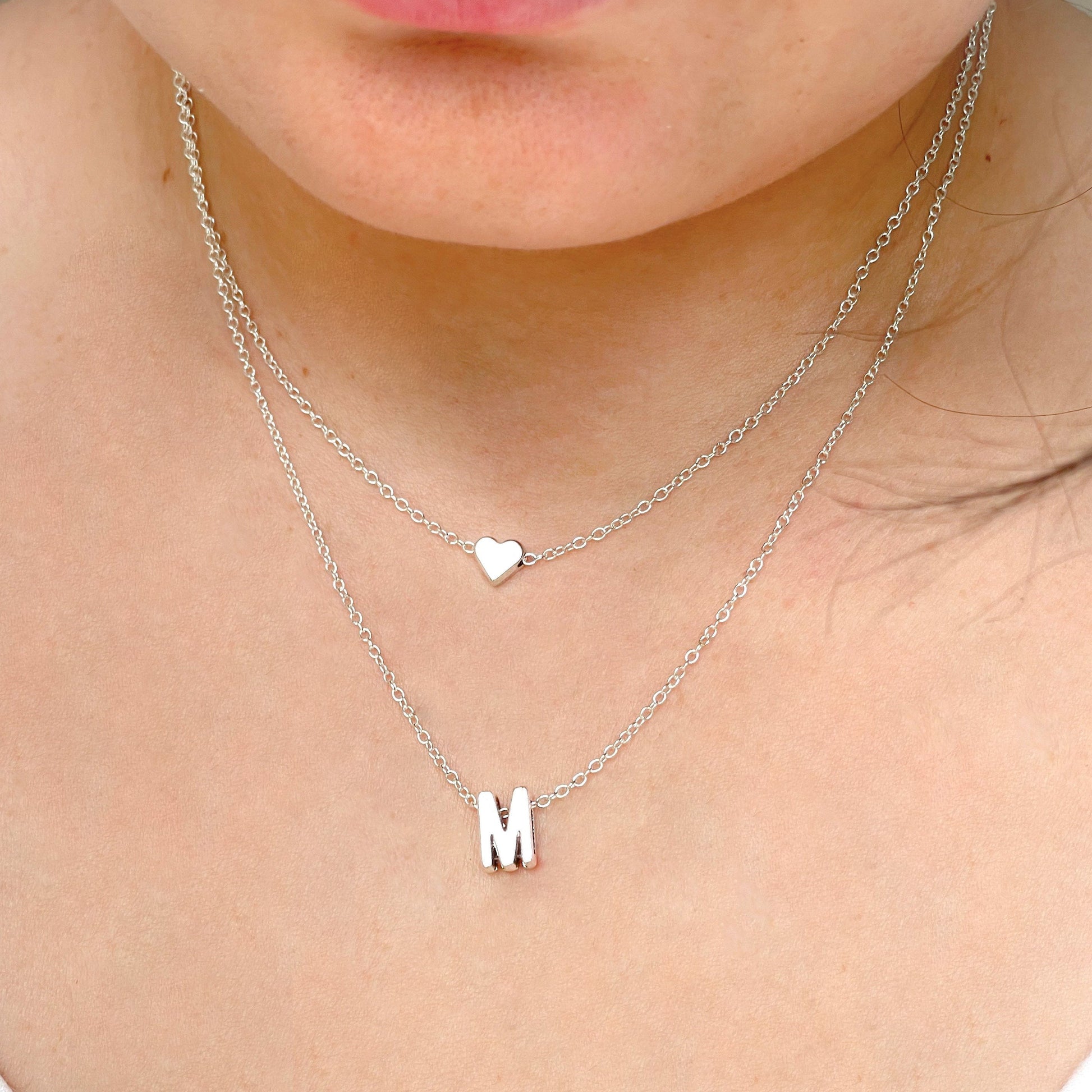 Layered silver necklace with heart and letter 'M' pendants on a person's neck.