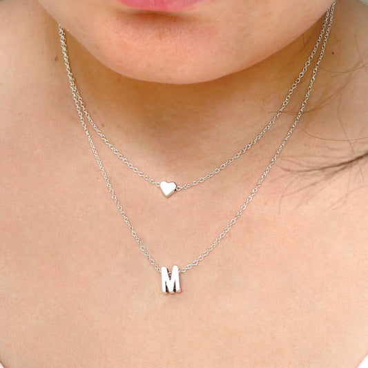 Layered silver necklace with heart and letter 'M' pendants on a person's neck.