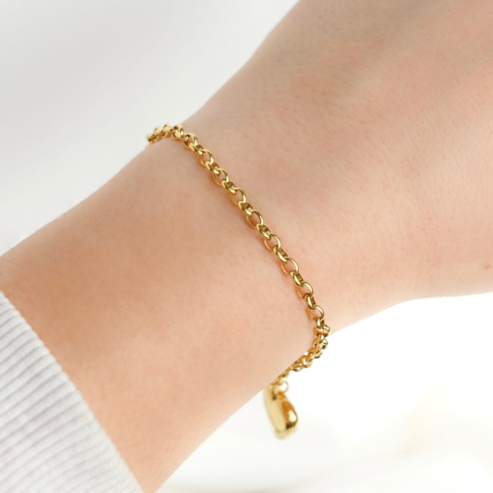 A delicate gold rolo chain bracelet with a big dangling heart charm, shown worn on a person's wrist against a bright white background.