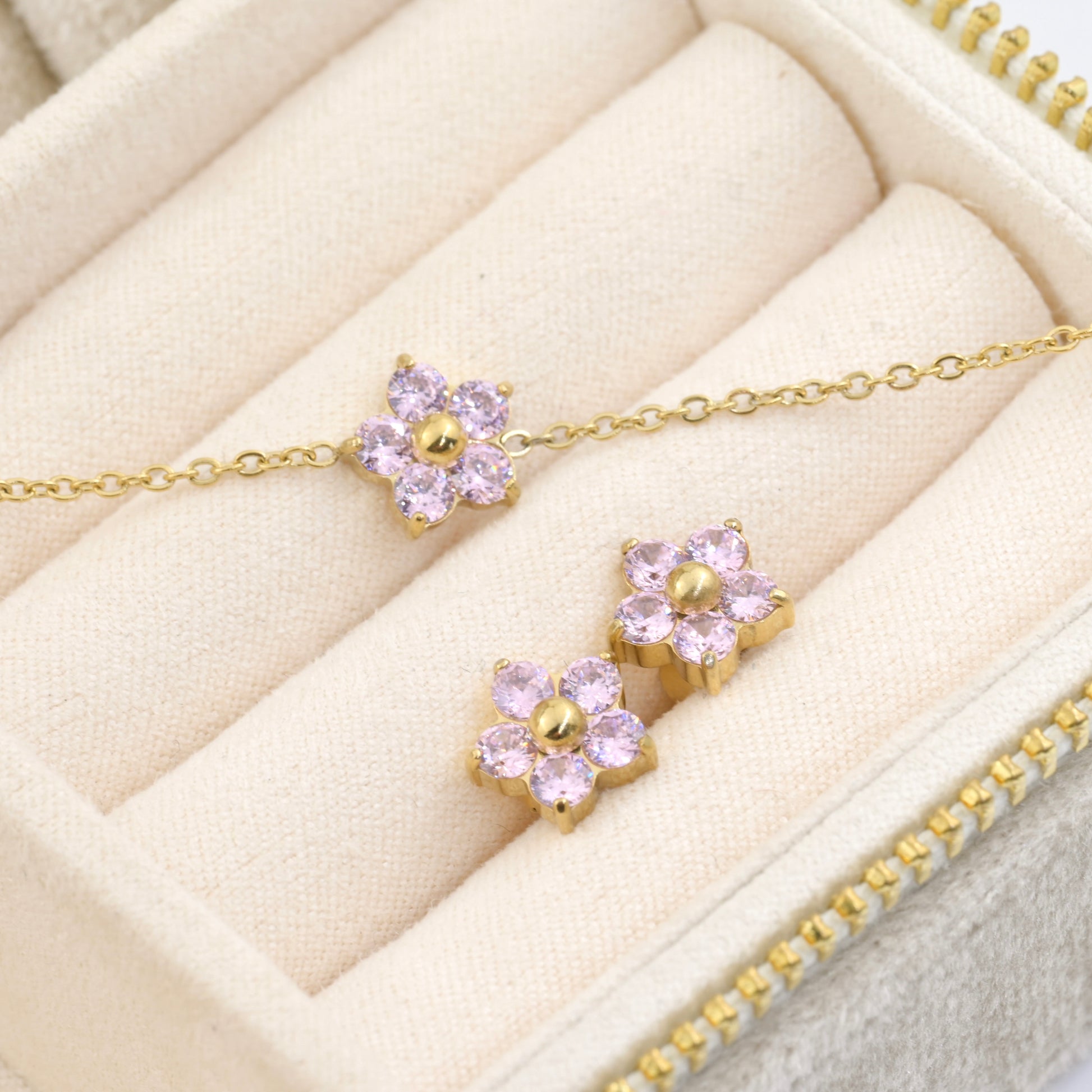 Gold bracelet and two stud earrings with pink CZ on a beige jewelry box.
