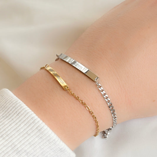 Two engraved bracelets, one gold with small bar and a cable, one silver with big bar and a dainty curb chain, on a wrist against a neutral background.