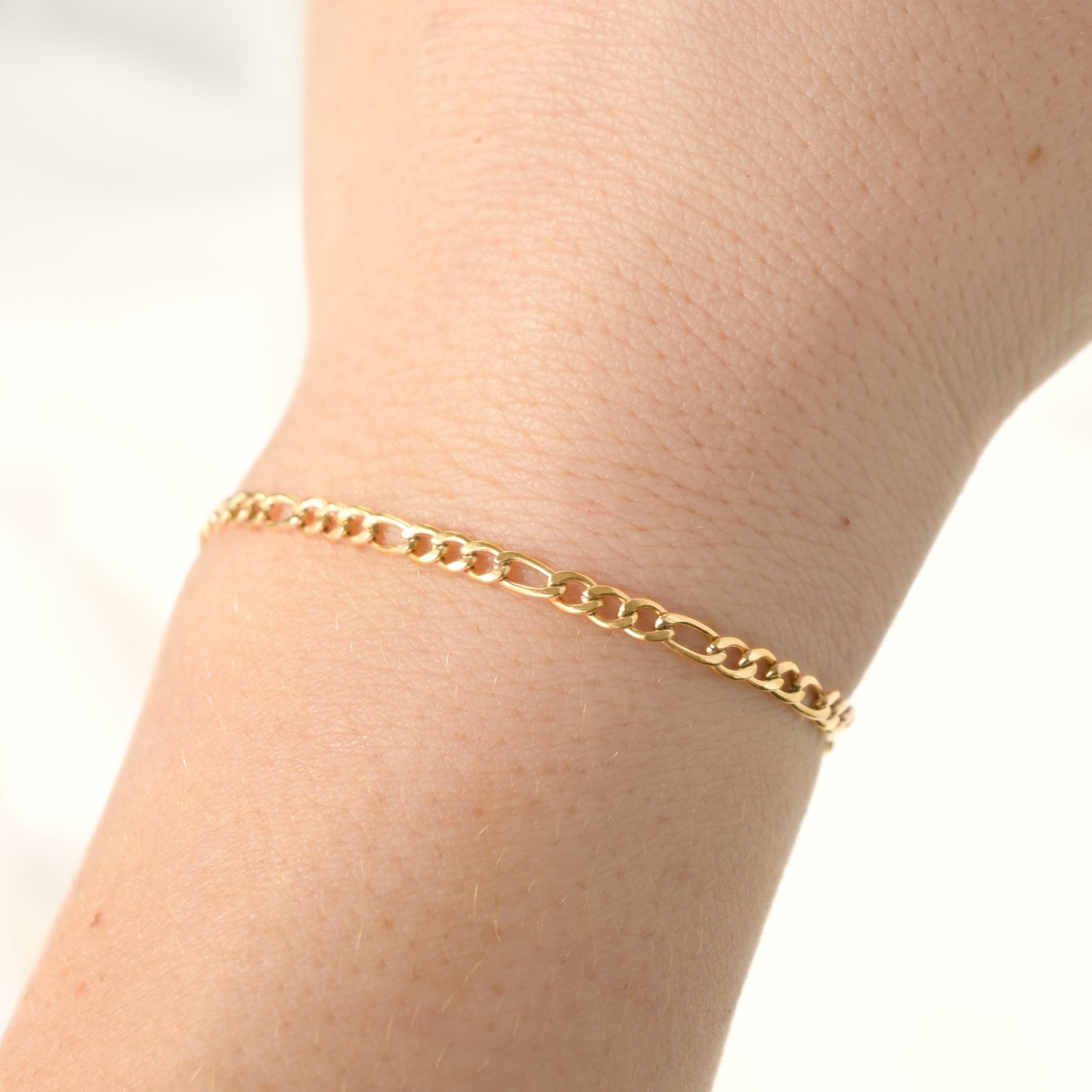 Gold figaro chain bracelet on a wrist with a plain background