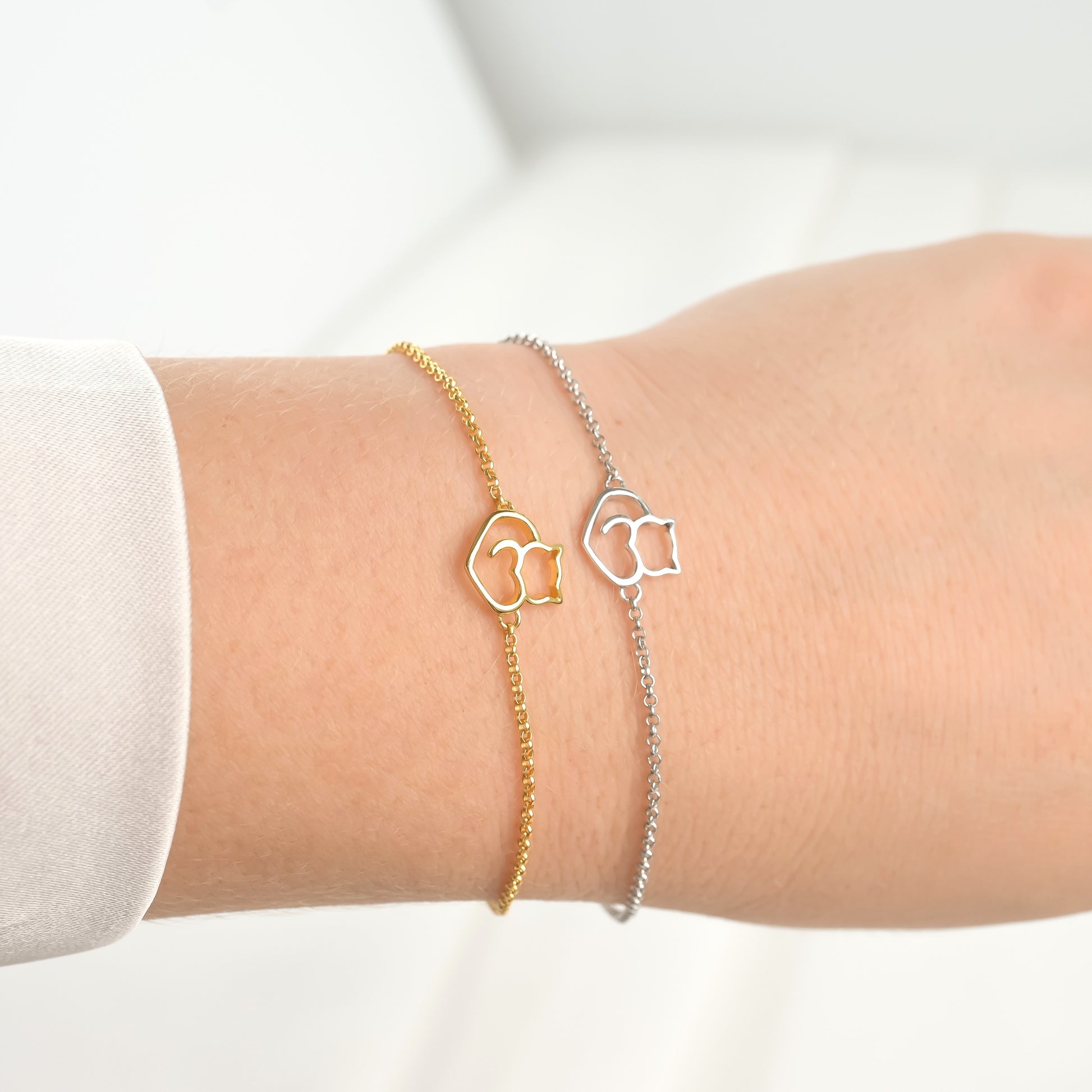 Two cat charm bracelets, one gold and one silver, on a wrist against a white background