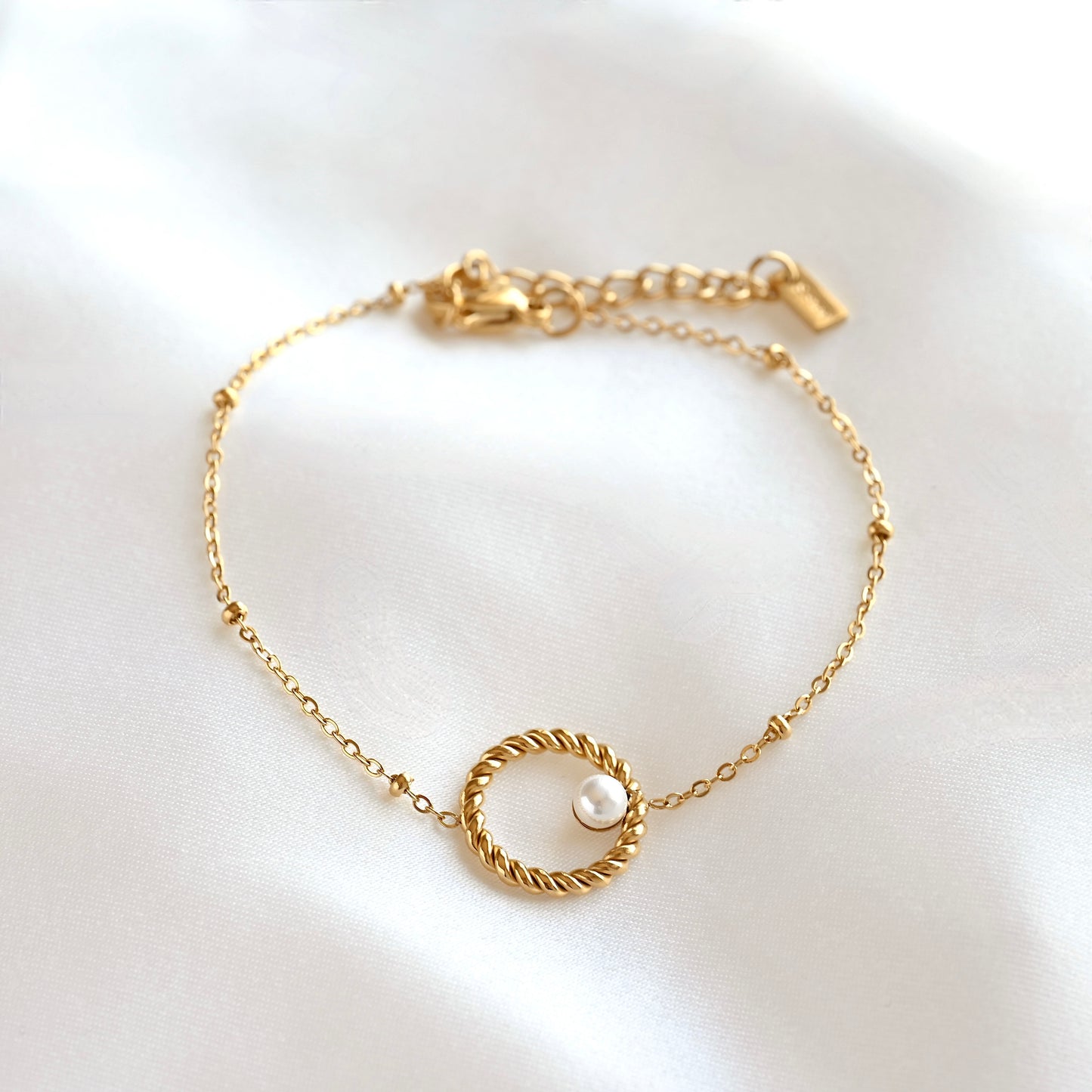 A delicate gold chain bracelet with a central twisted rope-style circle and a small white pearl, displayed on a soft white satin fabric background.