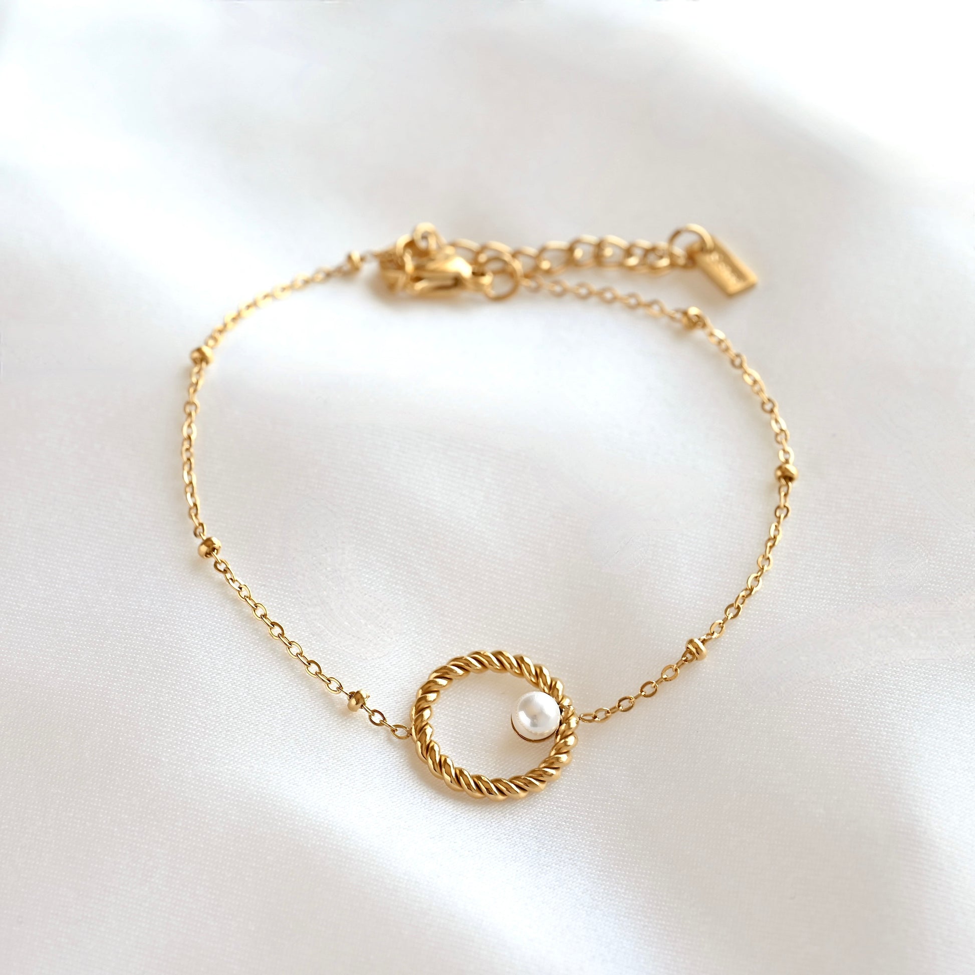 A delicate gold chain bracelet with a central twisted rope-style circle and a small white pearl, displayed on a soft white satin fabric background.