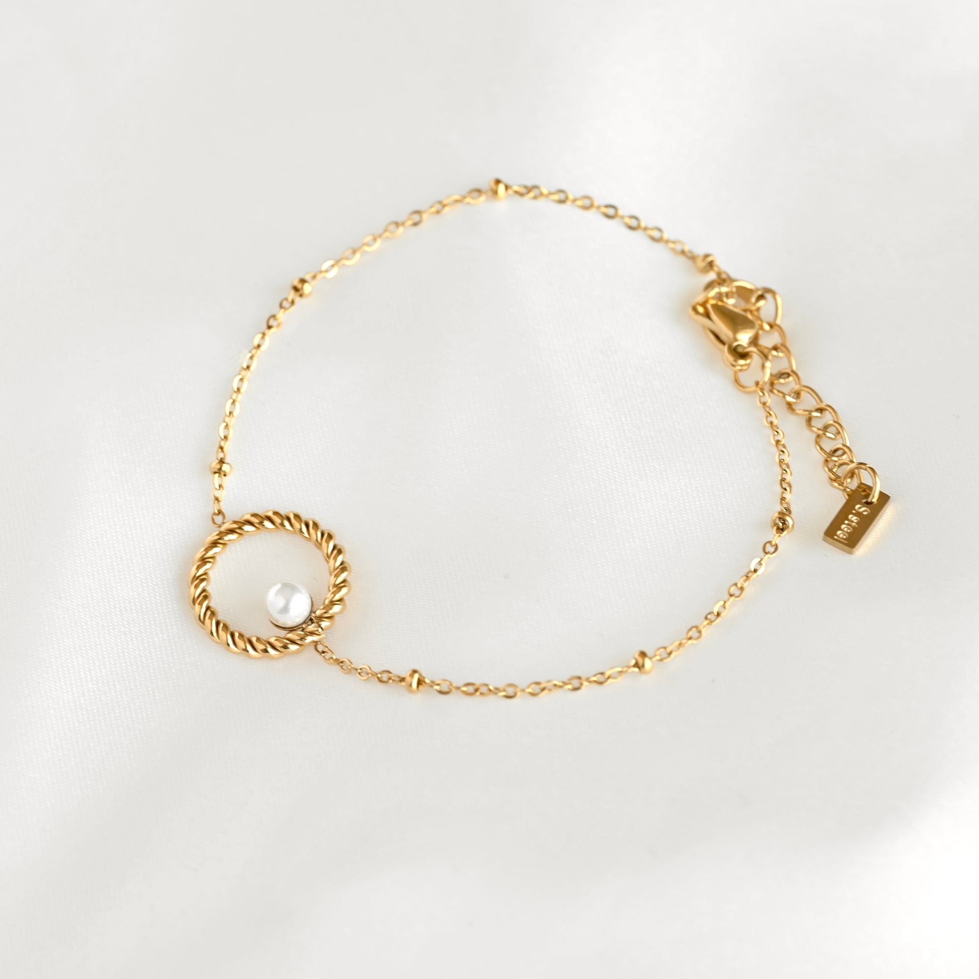 A delicate gold chain bracelet with a twisted circular ring holding a small white pearl, displayed on a soft white fabric background.