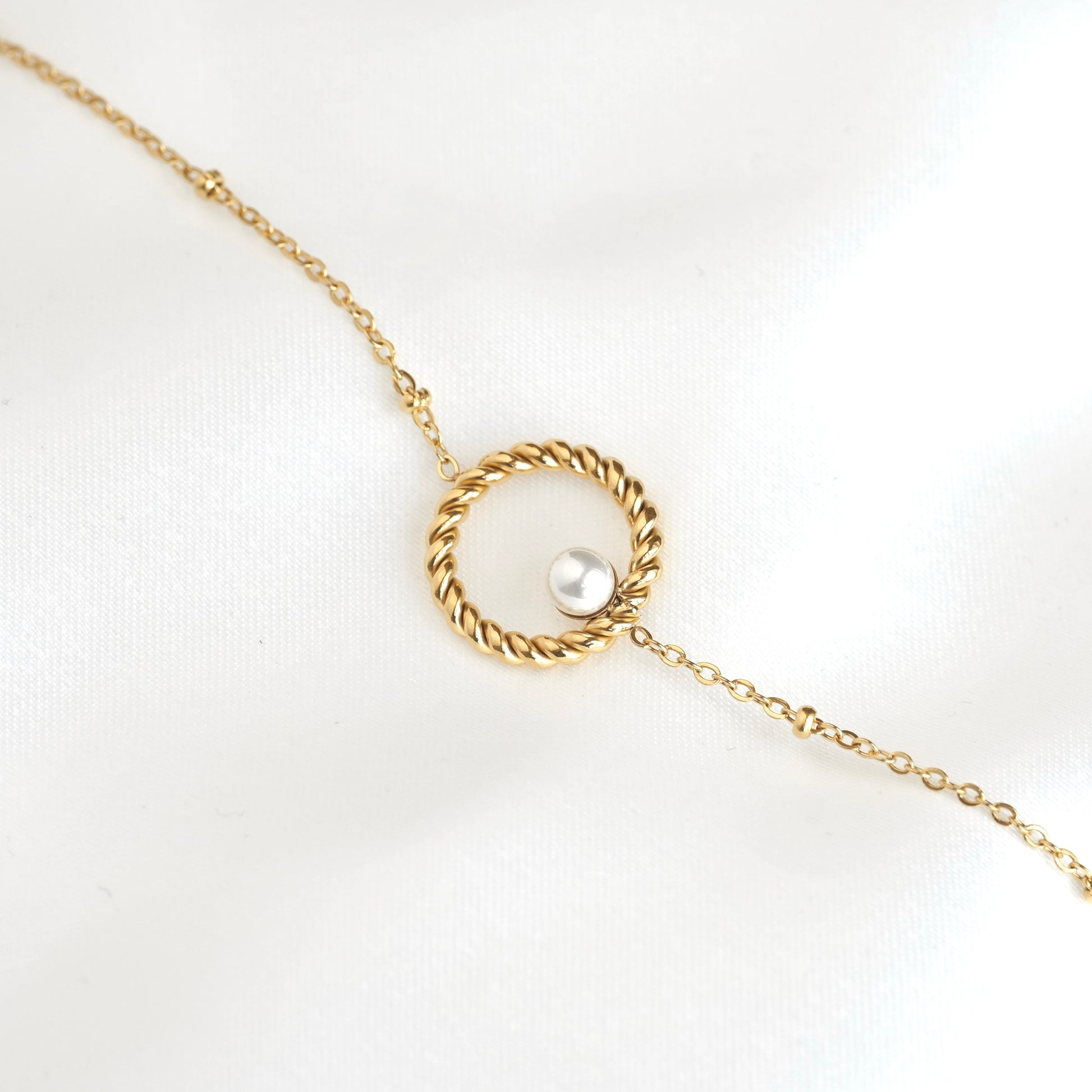 A delicate gold chain bracelet featuring a twisted rope-style circular pendant with a single white pearl set in the center, displayed on a soft white fabric background.