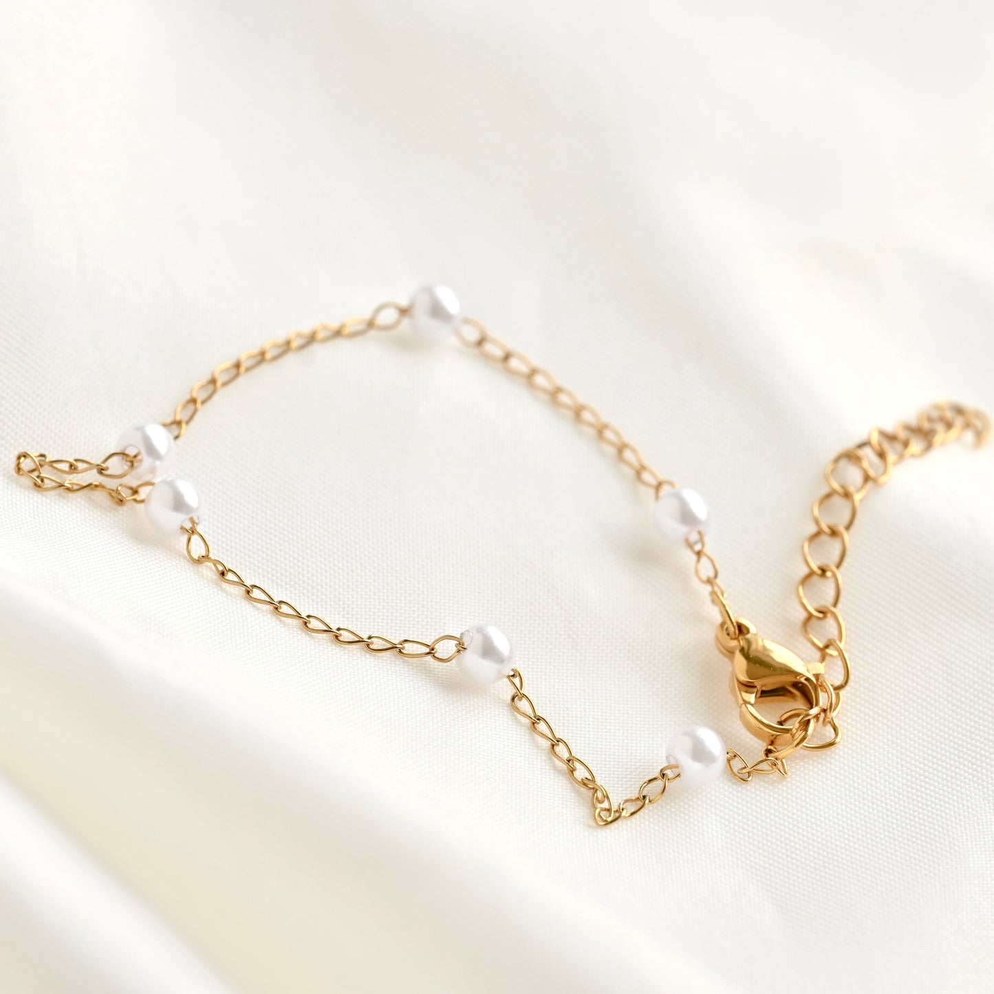 A delicate gold chain bracelet with small white pearl-like beads, displayed on a soft, draped white fabric background.