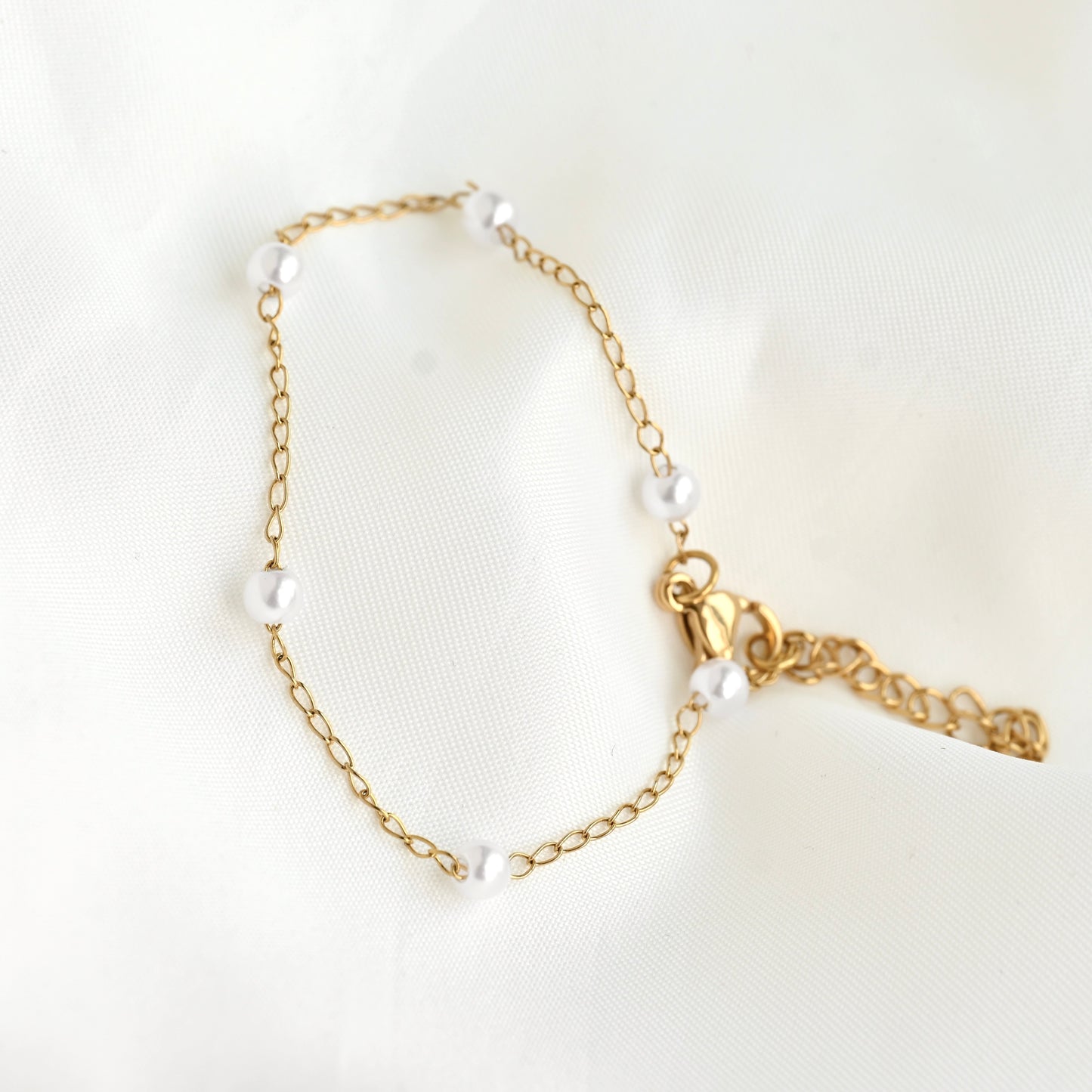 A delicate gold chain bracelet adorned with small white freshwater pearls, featuring a lobster clasp and an adjustable extender chain, displayed on a soft white fabric background.