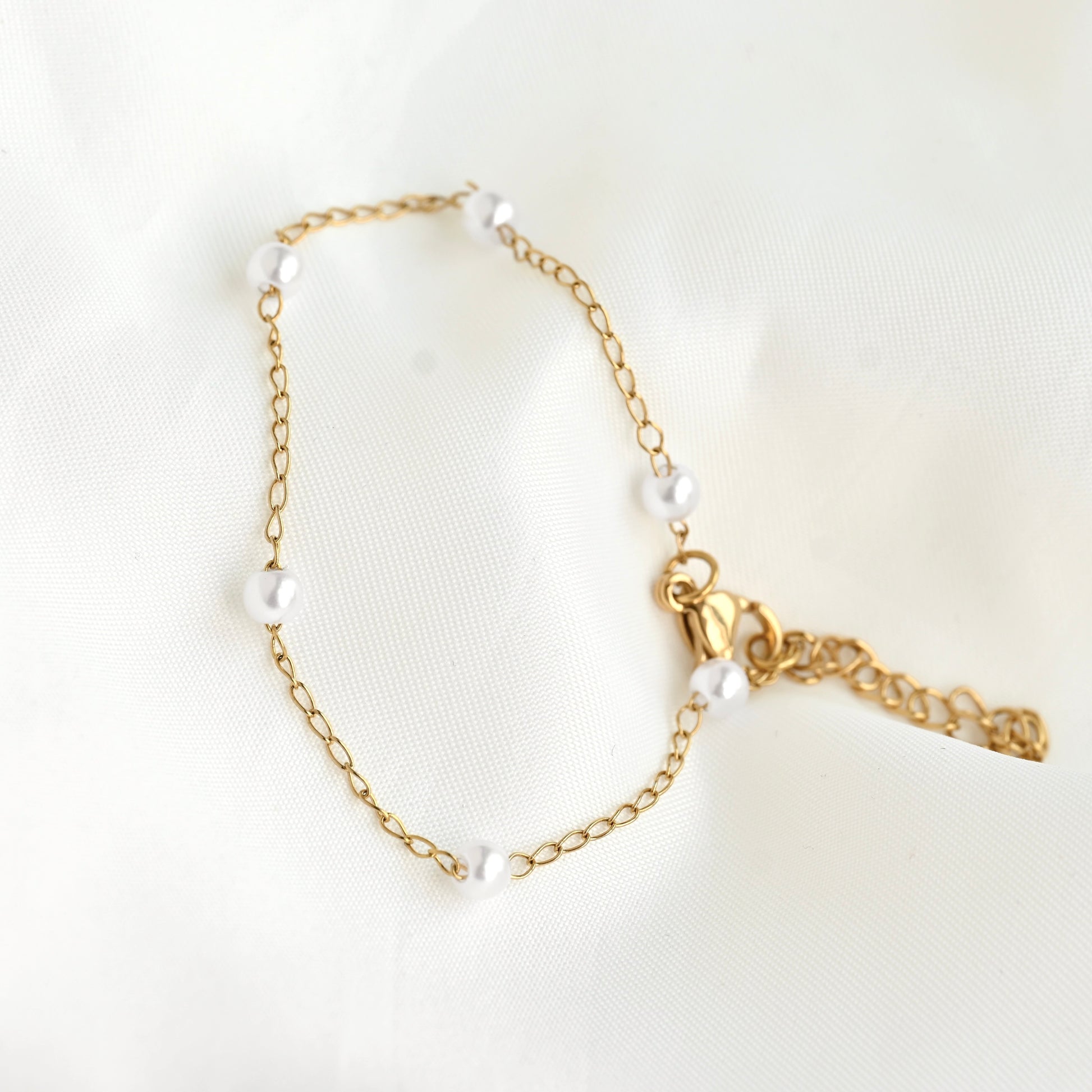 A delicate gold chain bracelet adorned with small white freshwater pearls, featuring a lobster clasp and an adjustable extender chain, displayed on a soft white fabric background.
