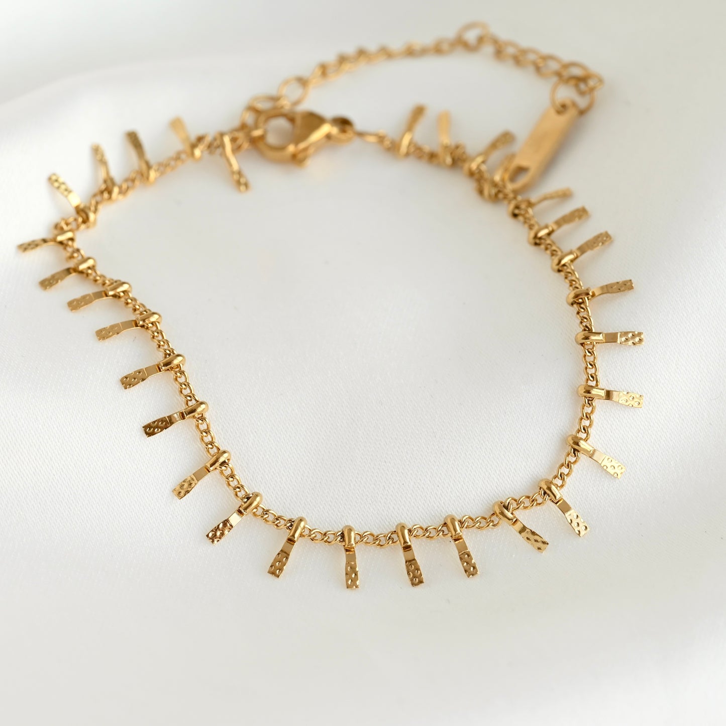 A gold chain bracelet with multiple textured, flat bar charms arranged in a zigzag pattern, displayed on a clean white background.