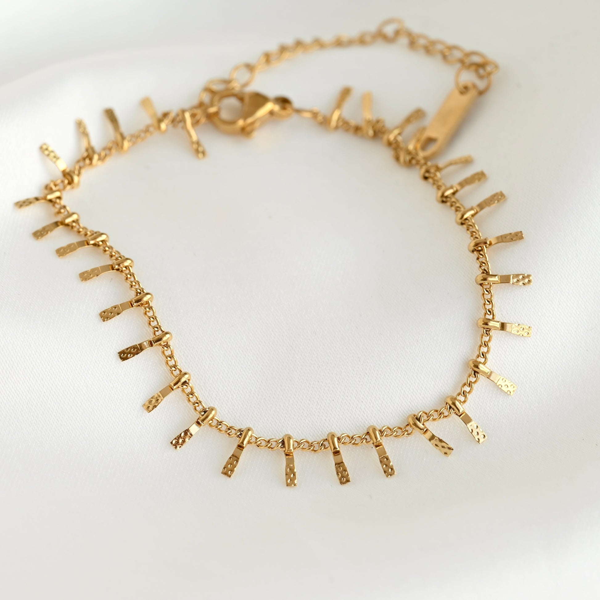 A gold chain bracelet with multiple textured, flat bar charms arranged in a zigzag pattern, displayed on a clean white background.