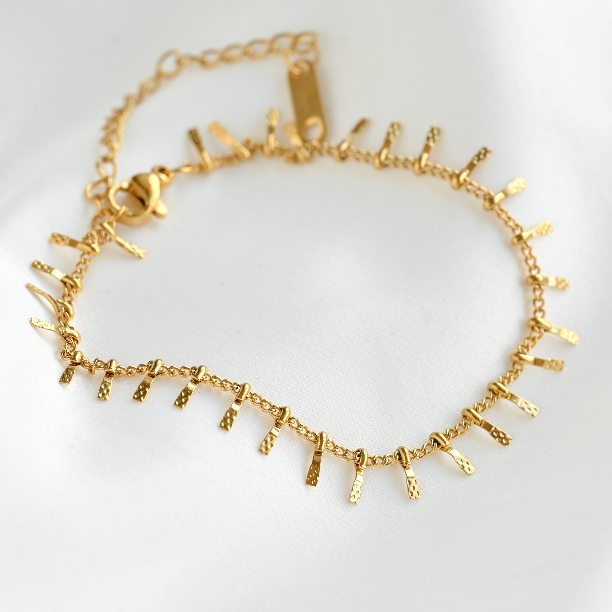 A delicate gold chain bracelet featuring small, textured vertical bar charms spaced along the front, displayed on a soft white fabric background.