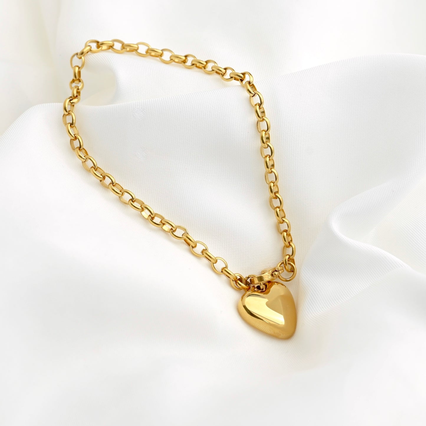 A gold rolo chain bracelet with a polished chunky heart-shaped charm, displayed on a soft white fabric background.