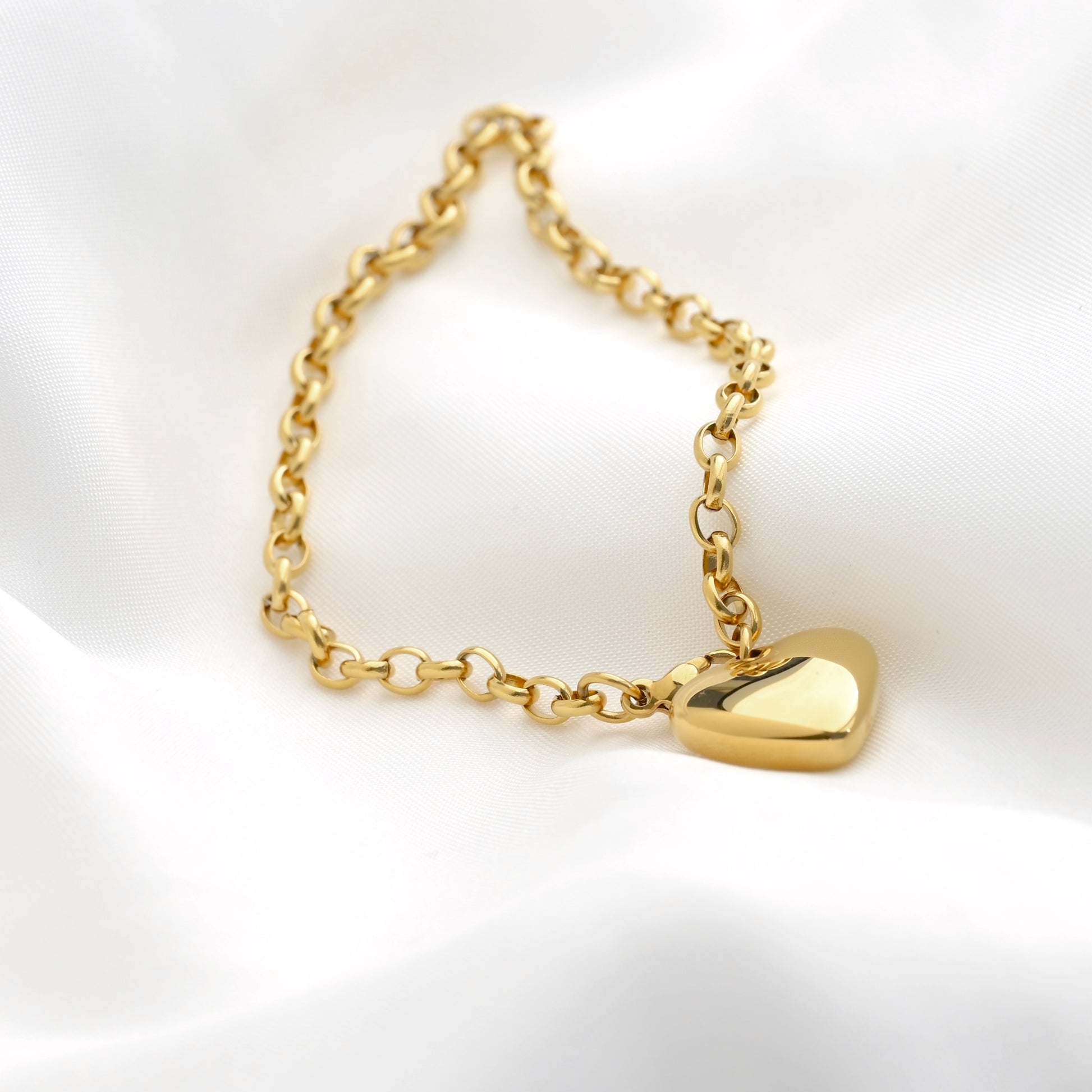 A gold chain bracelet with a polished heart-shaped pendant, displayed on a soft white satin fabric background.