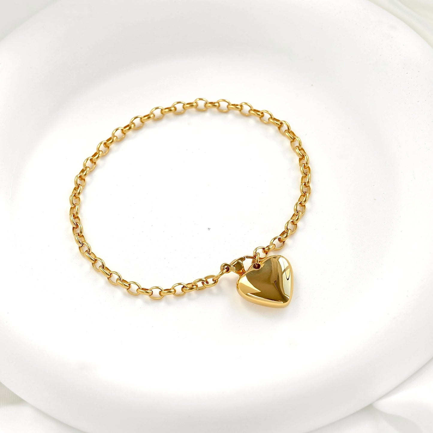 A delicate gold chain bracelet with a large, polished gold heart charm, displayed on a clean white surface.