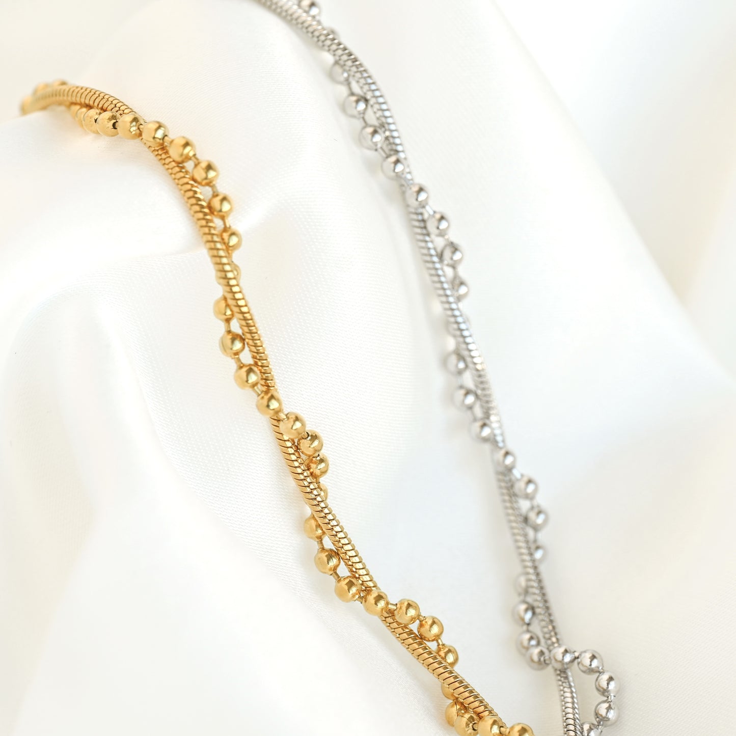 Two delicate chain bracelets draped on white satin fabric, one in polished yellow gold and one in polished silver, both featuring textured beaded links entwined with a thin snake chain.