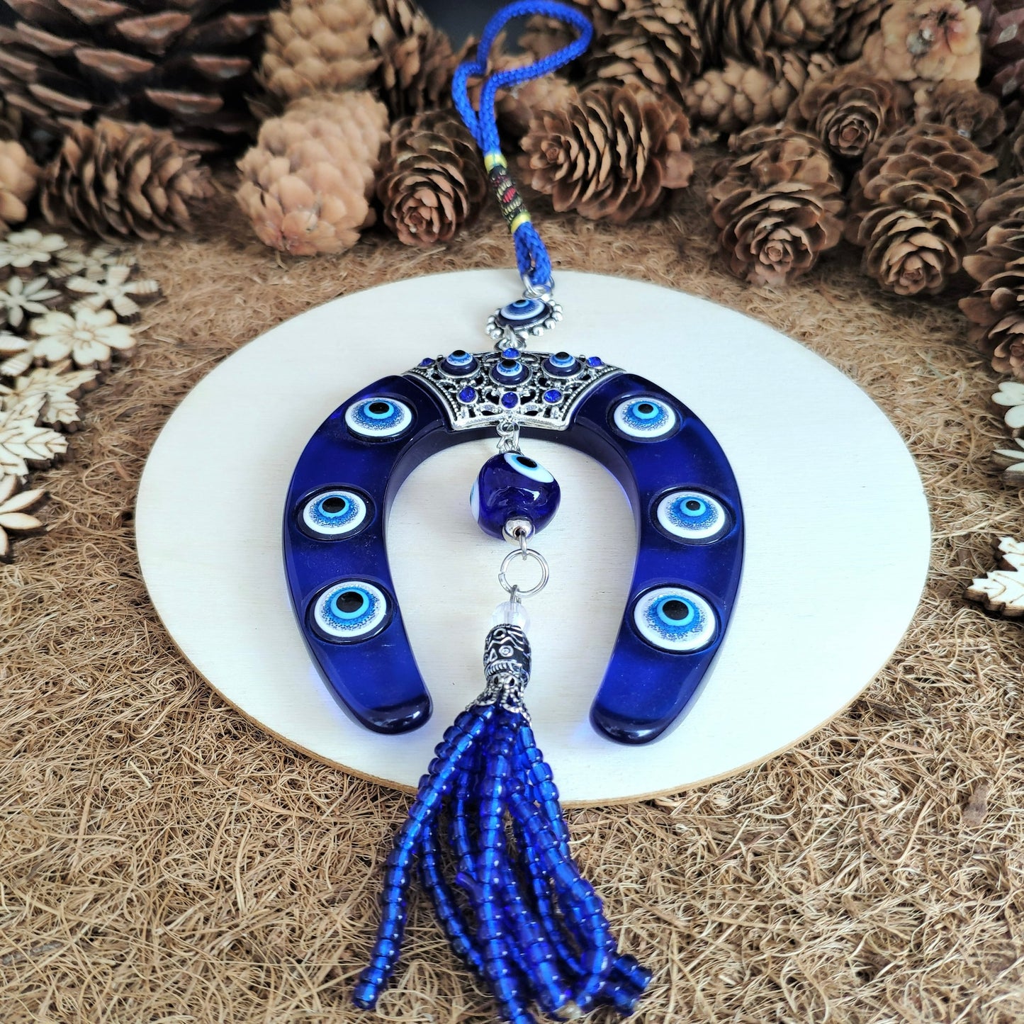 Close-up view of blue evil eye amulet with tassel on a white plate surrounded by pine cones and decorative elements.