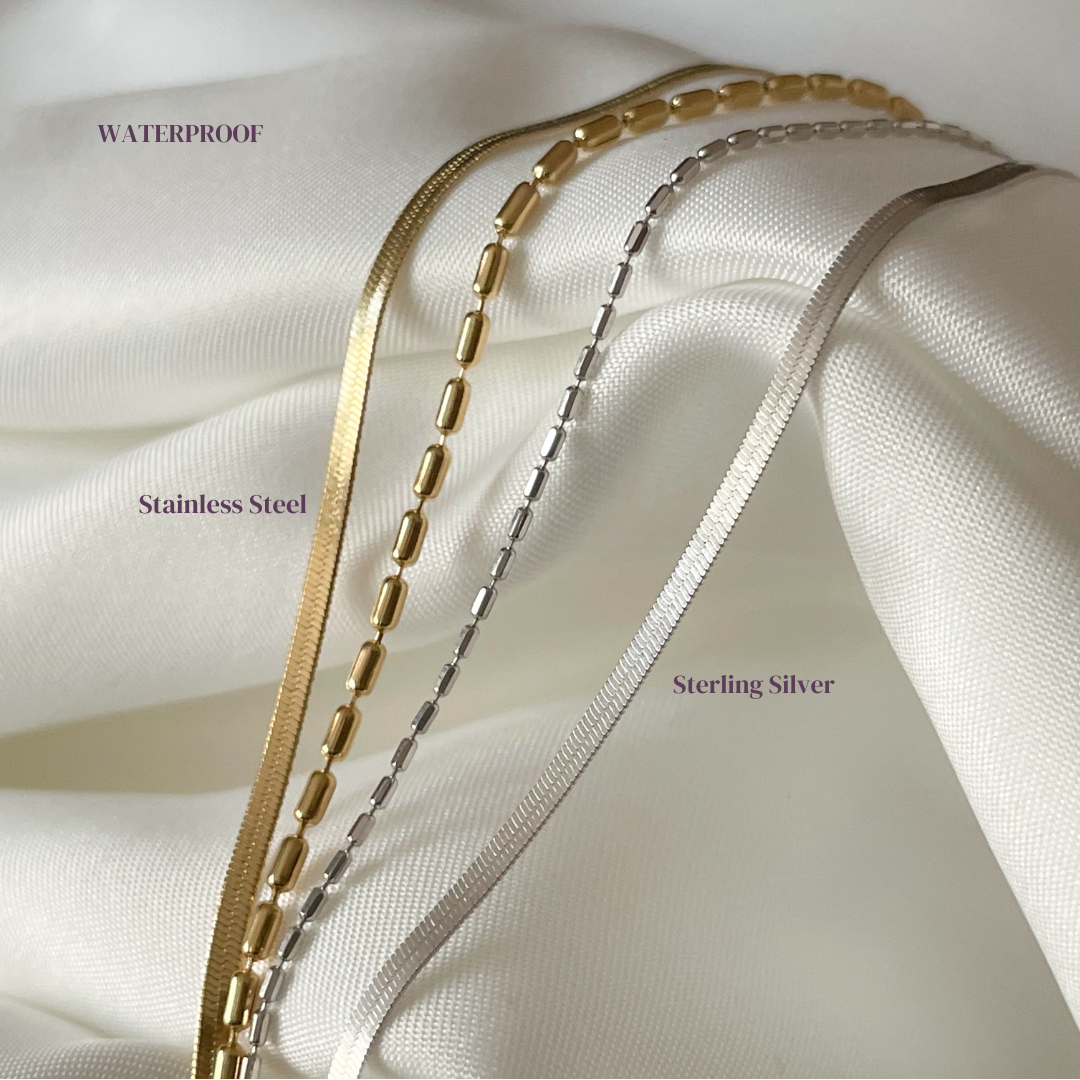 Two layered herringbone bracelets, one gold and one silver, on a white fabric background with stainless steel and sterling silver labels.