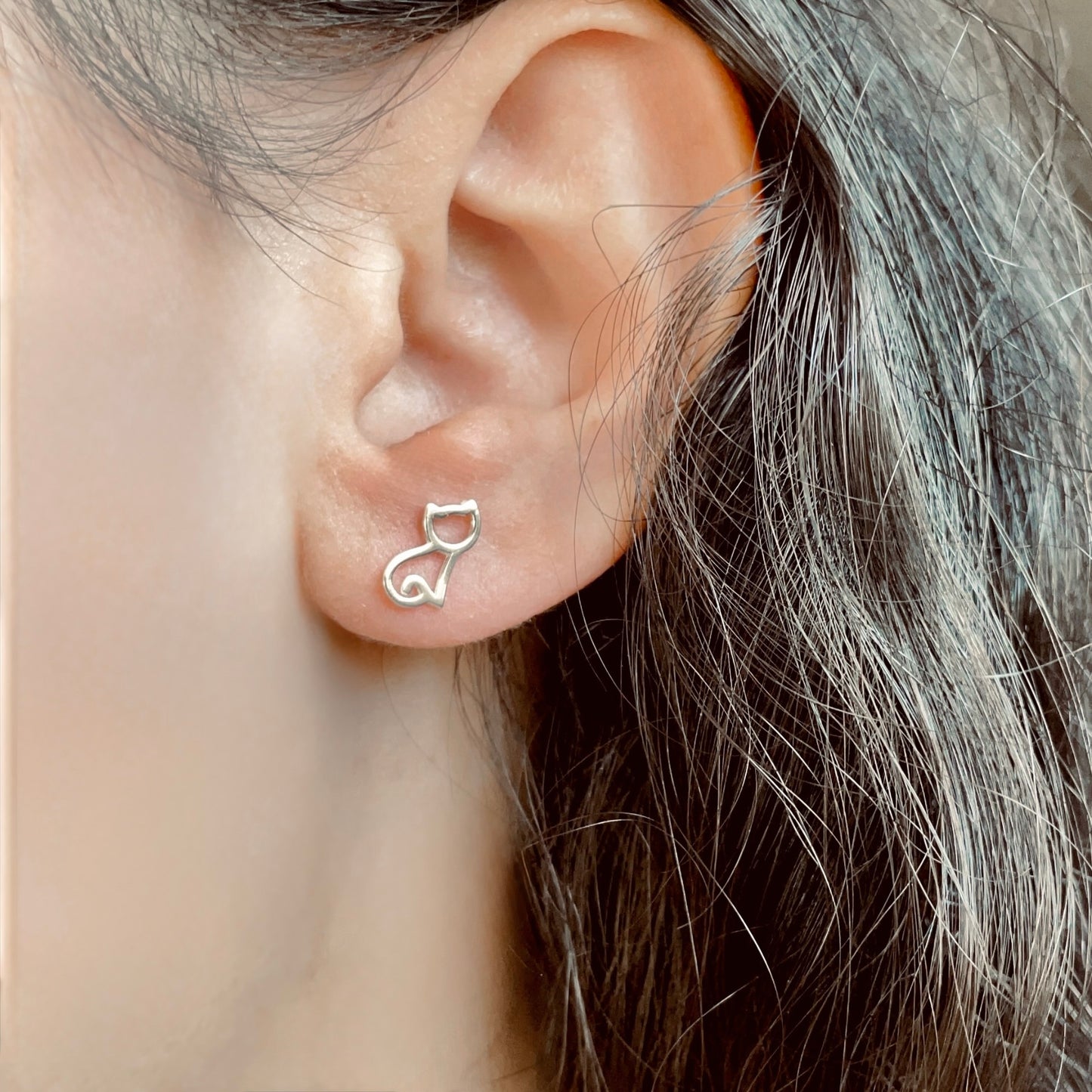Close-up of an ear wearing silver cat outline earrings with a blurred background