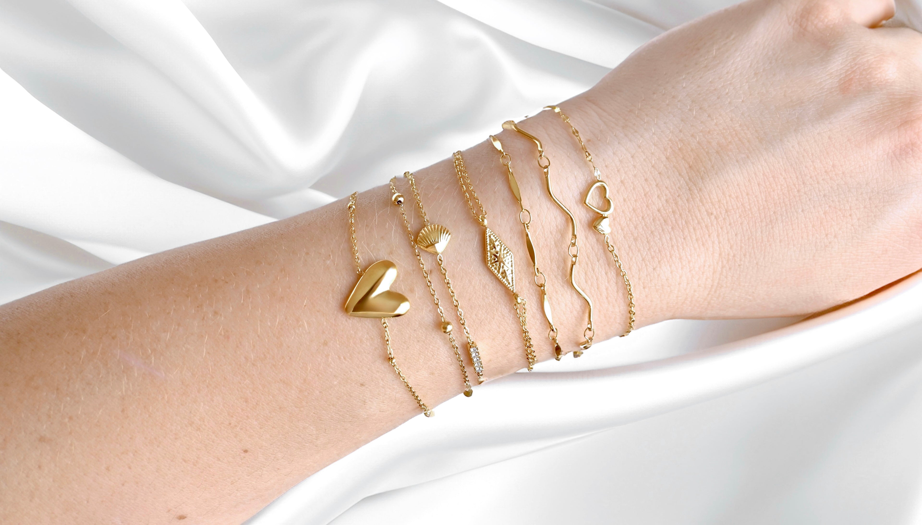 A gold bracelet stack on wrist against white satin fabric, the stack inclusing a large heart charm, a double layered seashell & cz bar, a double layered rhombus shaped sunburst charm, oval link, S-shaped noodle link, and a super dainty double heart bracelet with mixed chain.