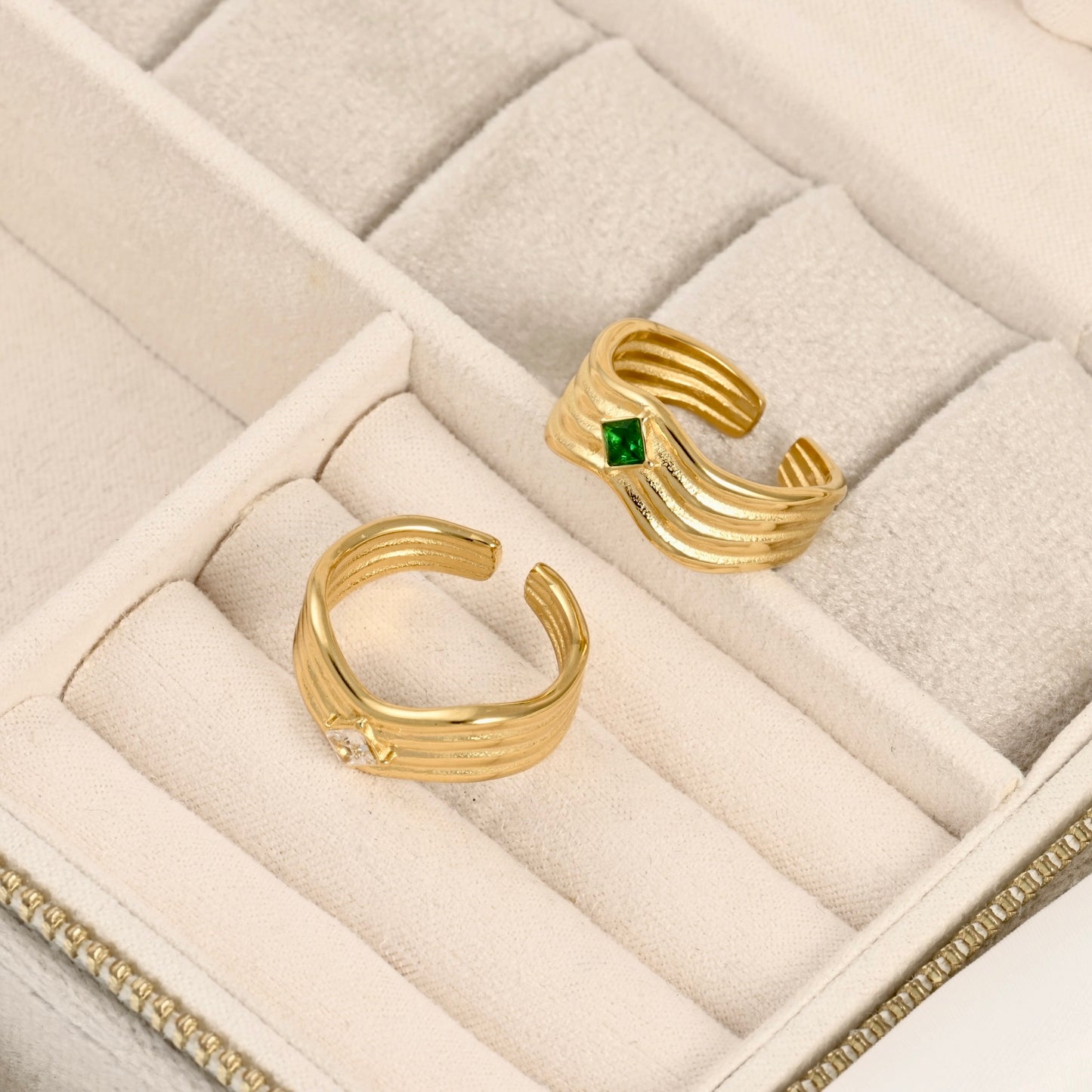 Two gold open-bend rings displayed in a cream-colored jewelry box. One ring features a small square green gemstone, and the other has a small clear round gemstone.