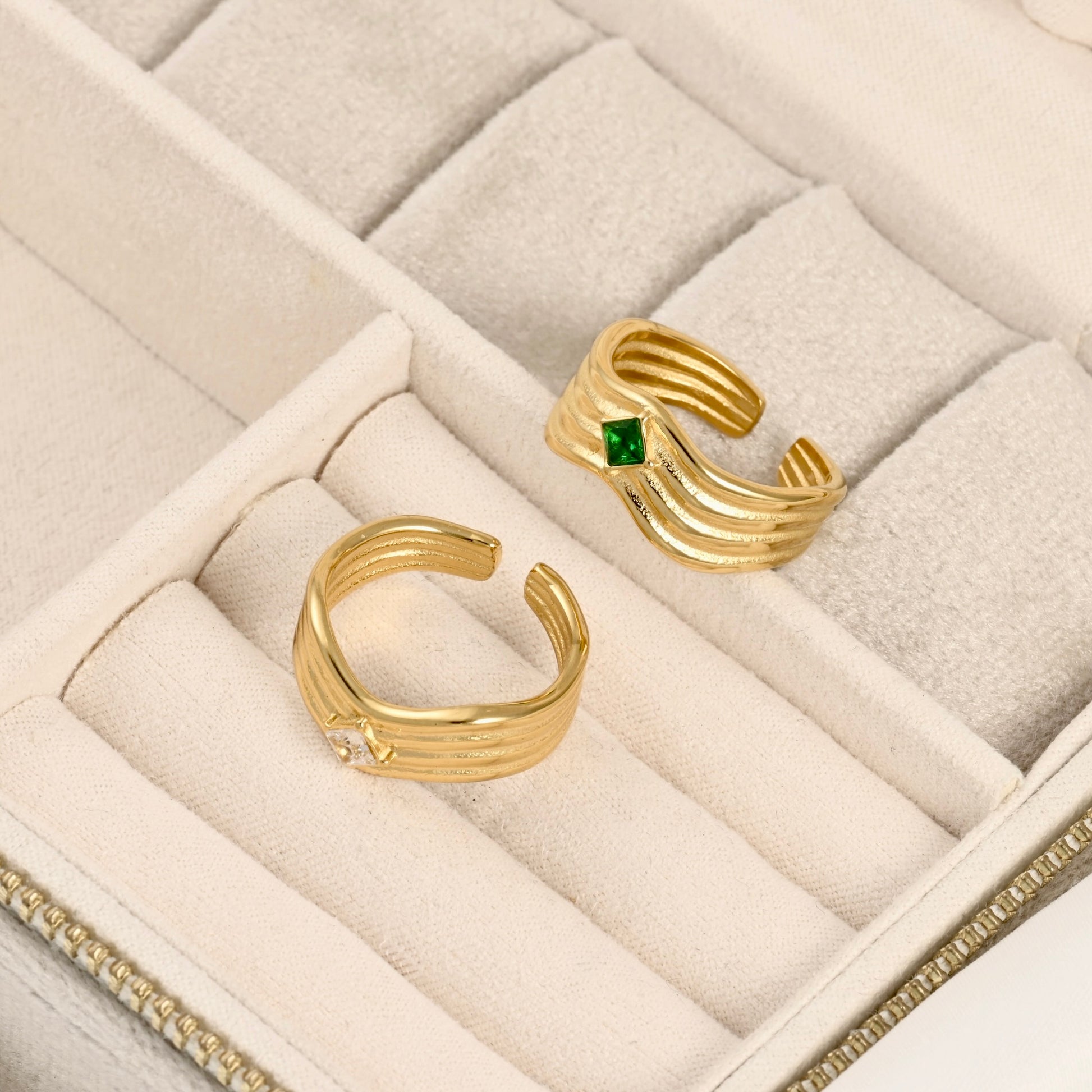 Two gold open-bend rings displayed in a cream-colored jewelry box. One ring features a small square green gemstone, and the other has a small clear round gemstone.