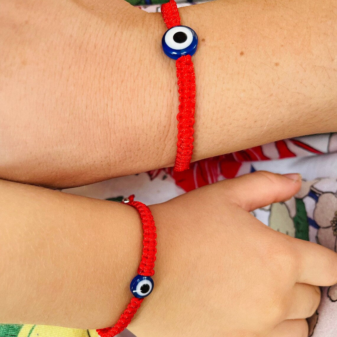 Red bracelets with blue evil eye beads on a child's and women's wrists against a floral background